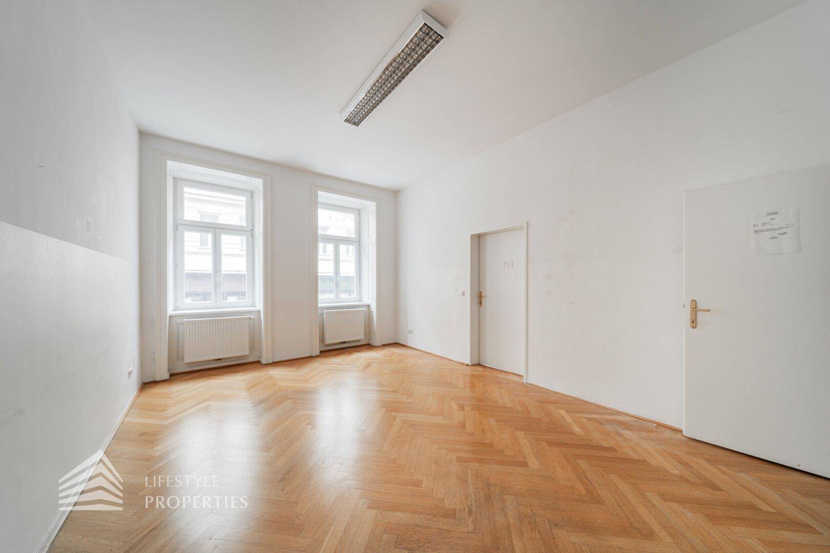 Großer Raum mit Fischgrätparkett und zwei Fenstern, ideal für ein geräumiges Büro oder Arbeitszimmer.