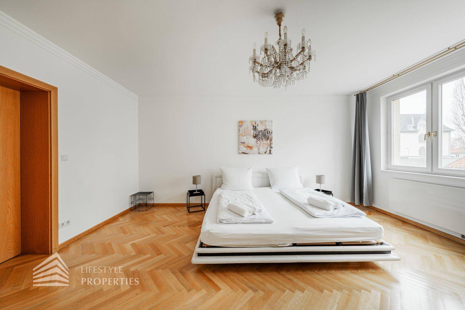Geräumiges Schlafzimmer mit Parkettboden, Kronleuchter und großem Fenster.