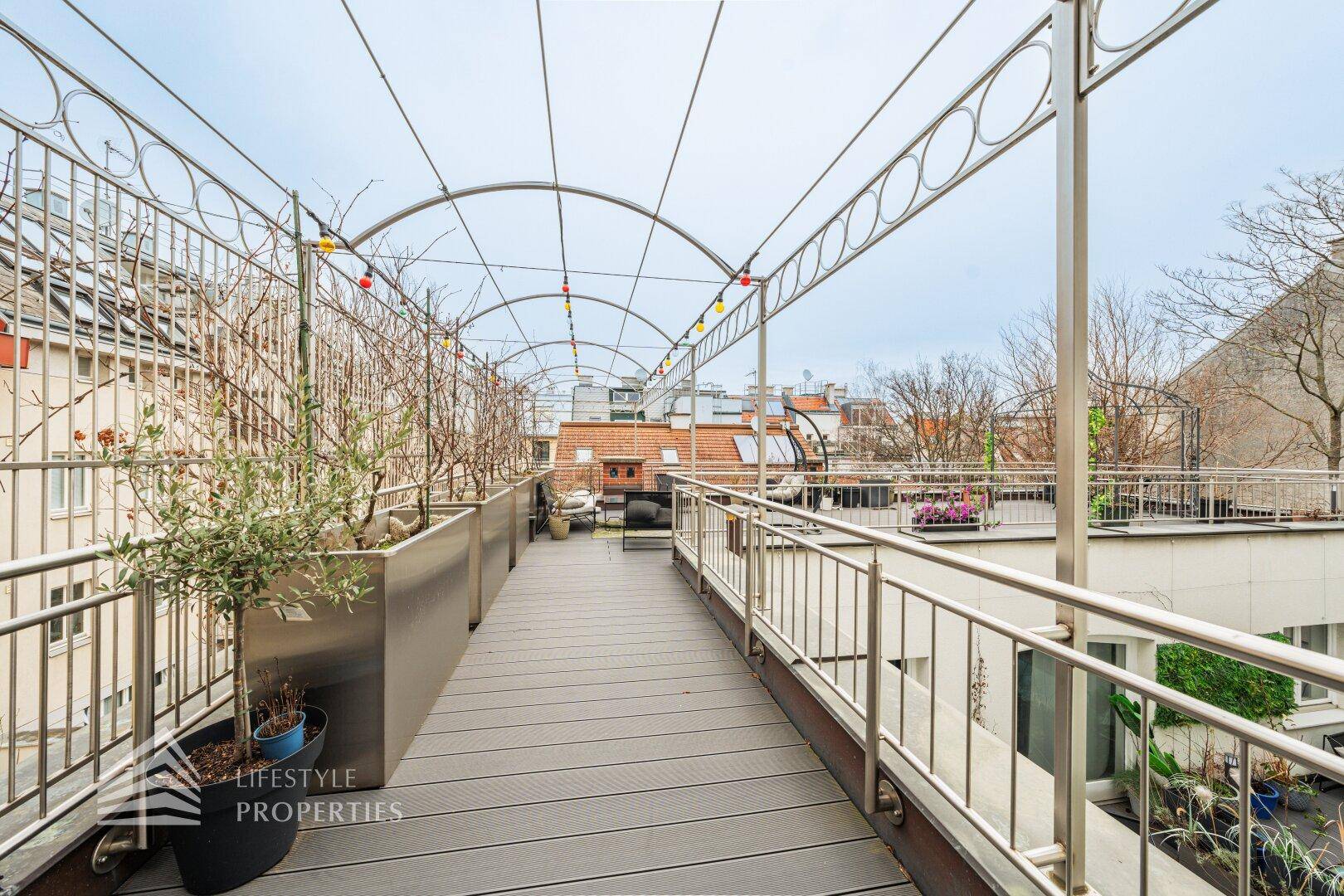 Lange Dachterrasse mit Metallpergola, Pflanzen und Blick über die Dächer.