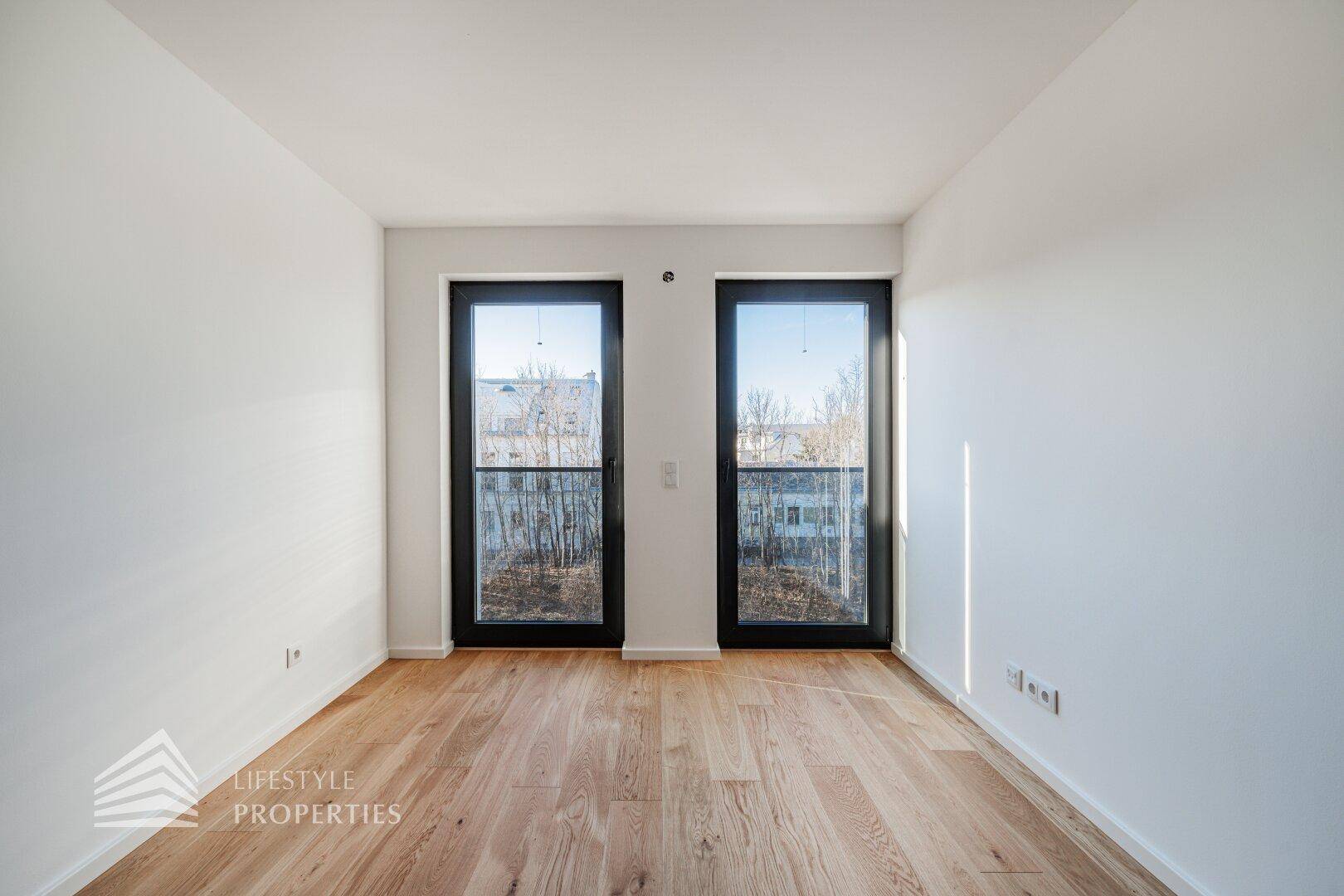 Lichtdurchfluteter Raum mit hellem Parkettboden und zwei hohen Fenstertüren, die den Blick nach draußen freigeben.