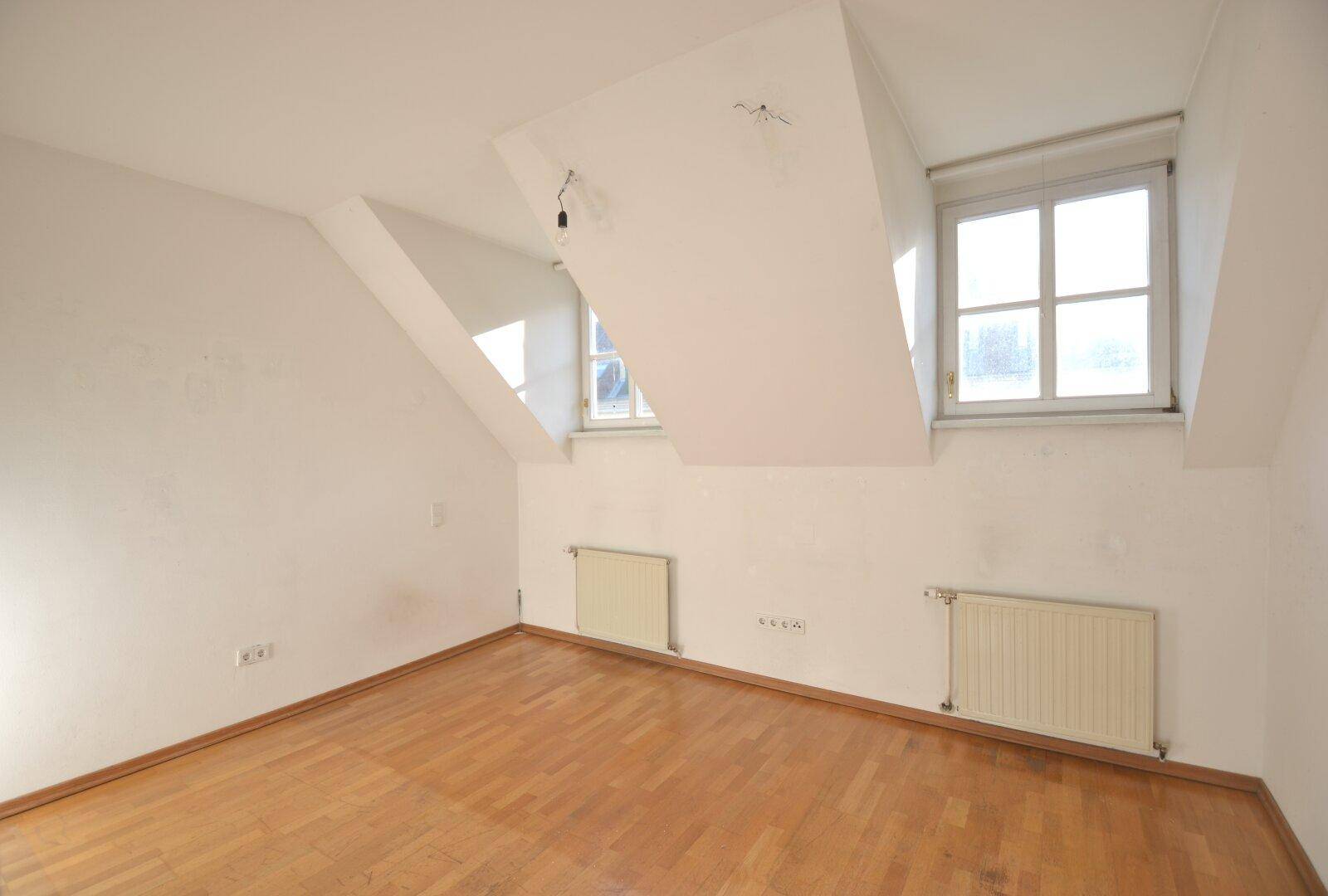 Leeres Schlafzimmer mit Holzboden, Dachschrägen und Fenstern, die natürliches Licht hereinlassen.