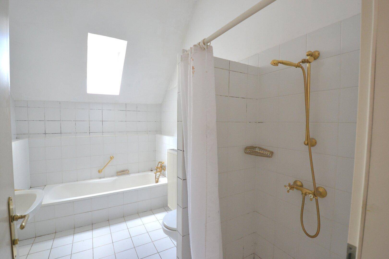 Helles Badezimmer mit Badewanne, Dusche und Dachfenster, ausgestattet mit goldenen Armaturen.