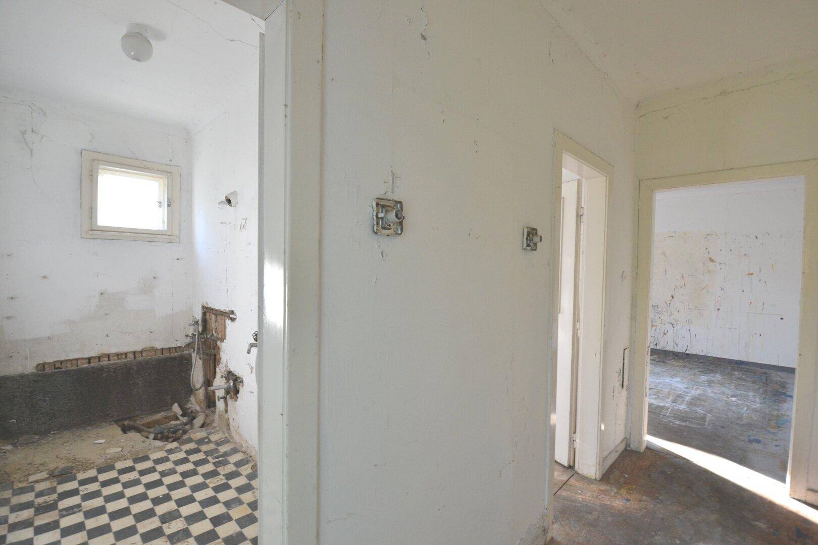 Flur mit Blick in ein sanierungsbedürftiges Badezimmer und einen weiteren leeren Raum.