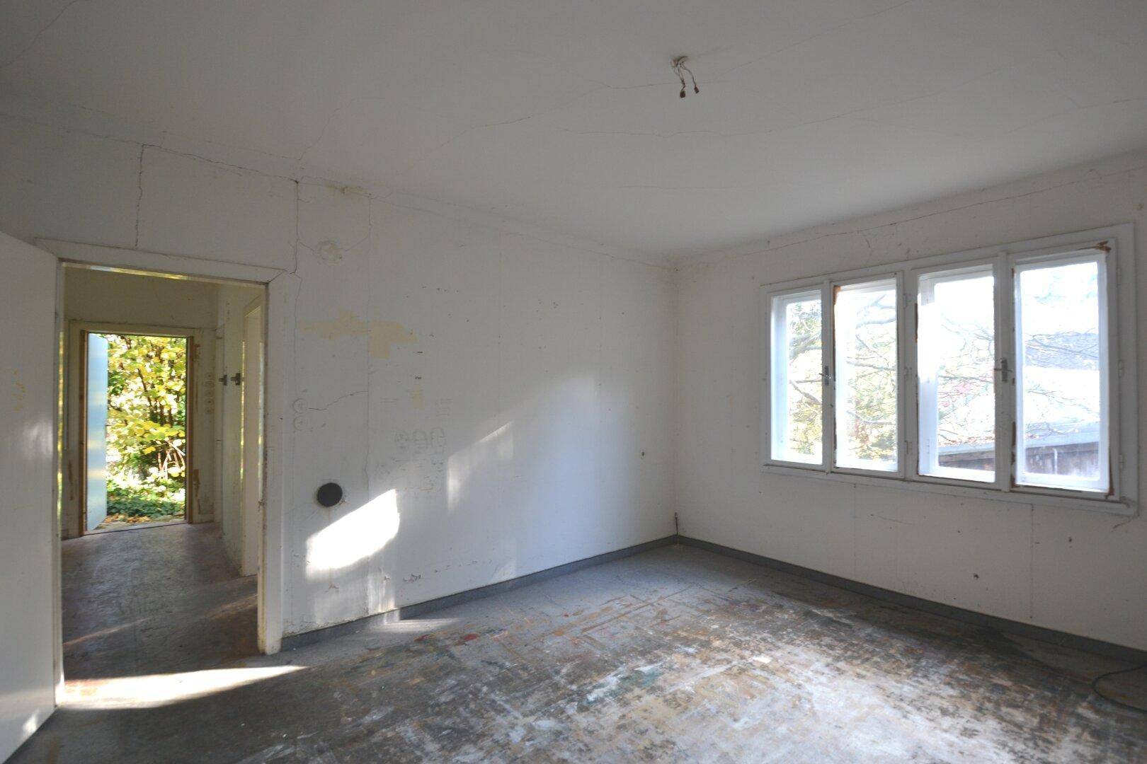 Leerer Raum mit abgenutztem Boden, weißen Wänden und Blick ins Grüne durch die Fenster.