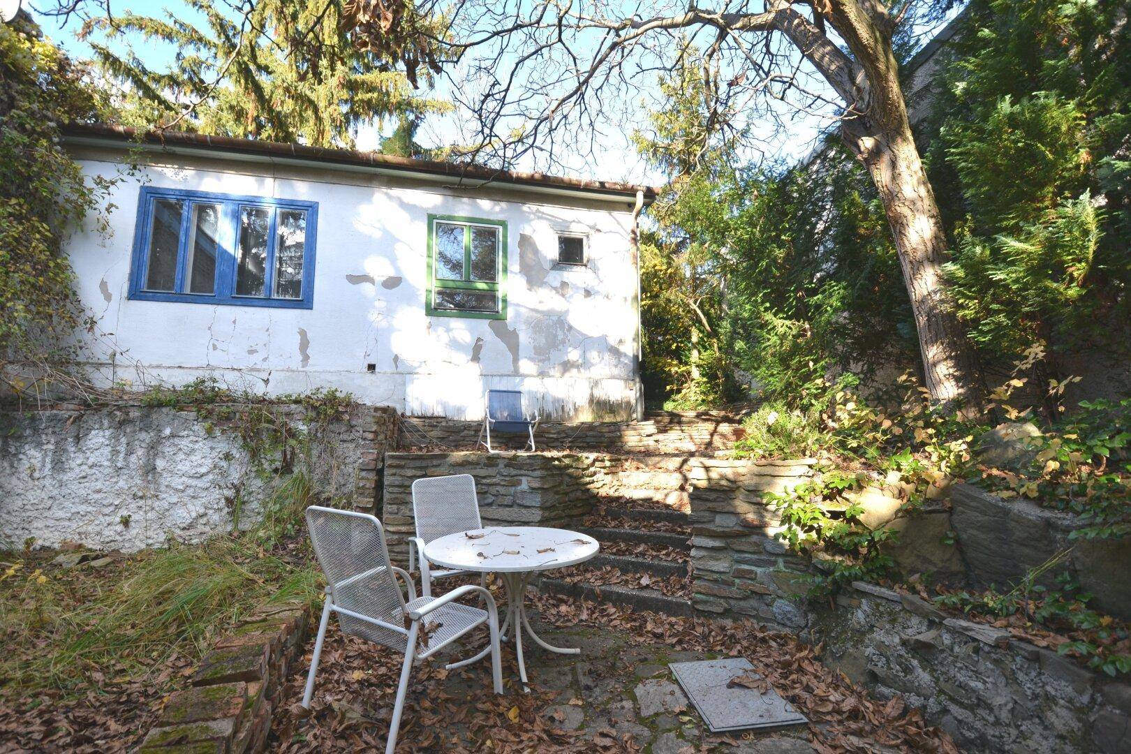 Kleine gepflasterte Terrasse mit Gartenmöbeln neben einem älteren Gebäude mit verputzten Wänden.