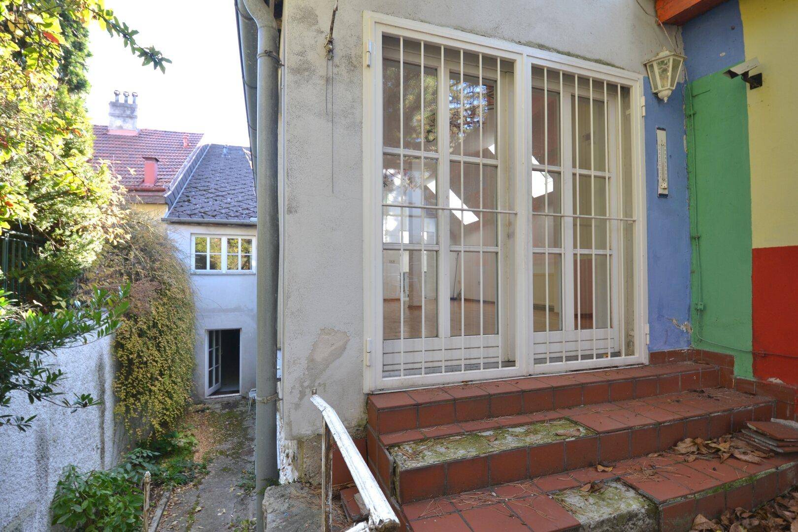 Außenansicht eines Gebäudes mit Balkon und Fenstergittern, die auf einen Gartenweg führen.