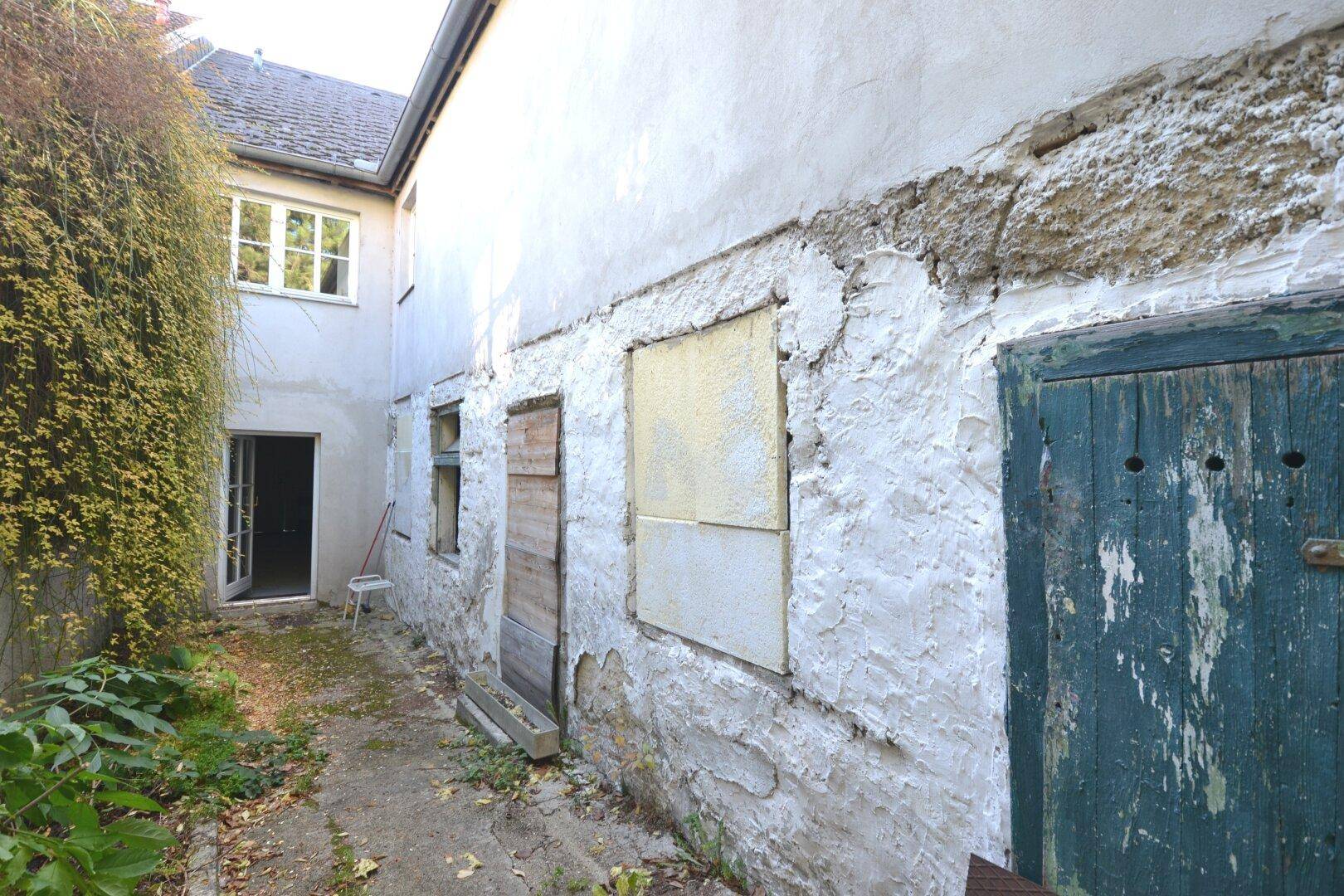 Seitenansicht eines alten Gebäudes mit verputzten Wänden, zugenagelten Fenstern und einem schmalen Durchgang.