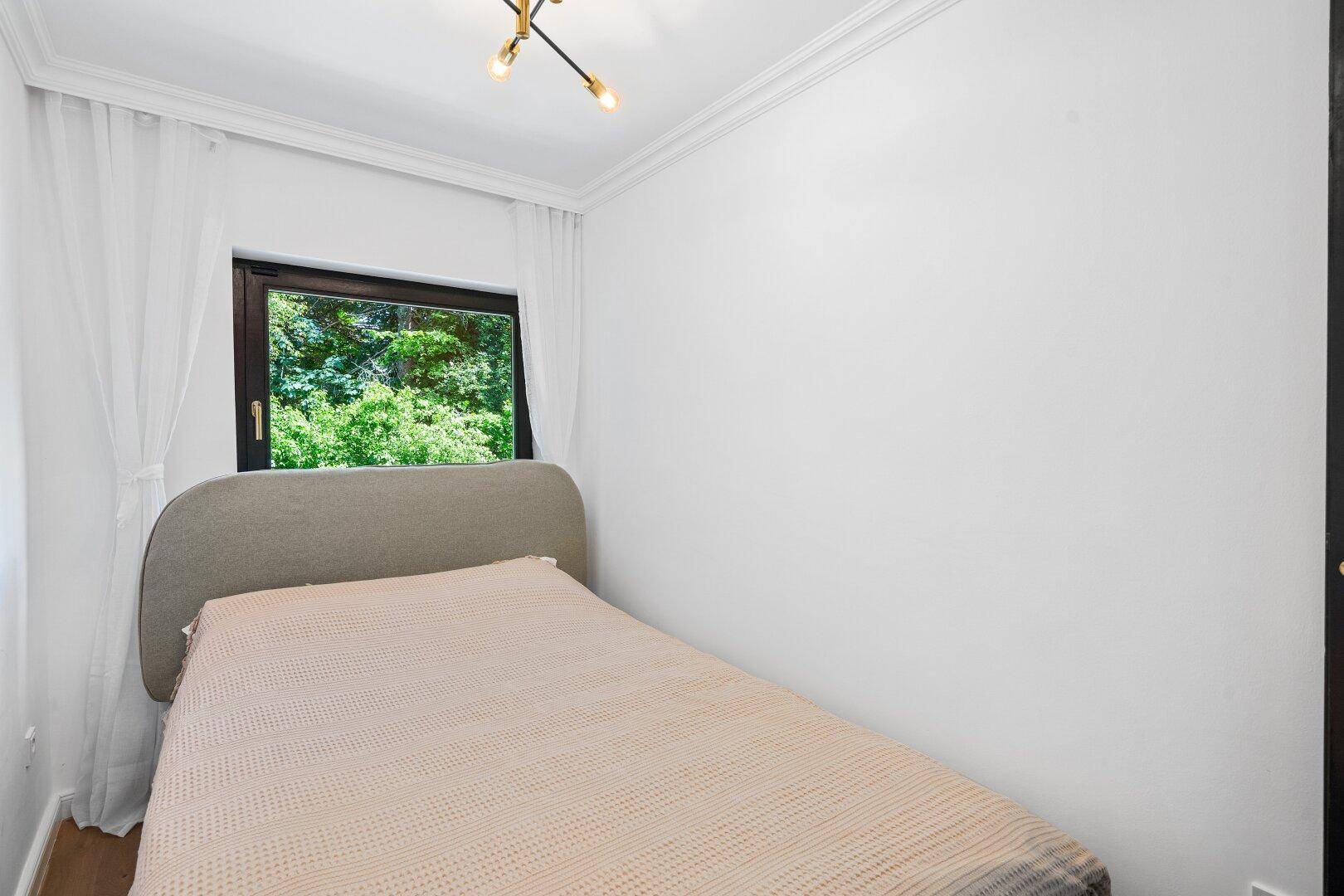Kleines Schlafzimmer mit Einzelbett, Holzboden und Fenster mit Blick ins Grüne.