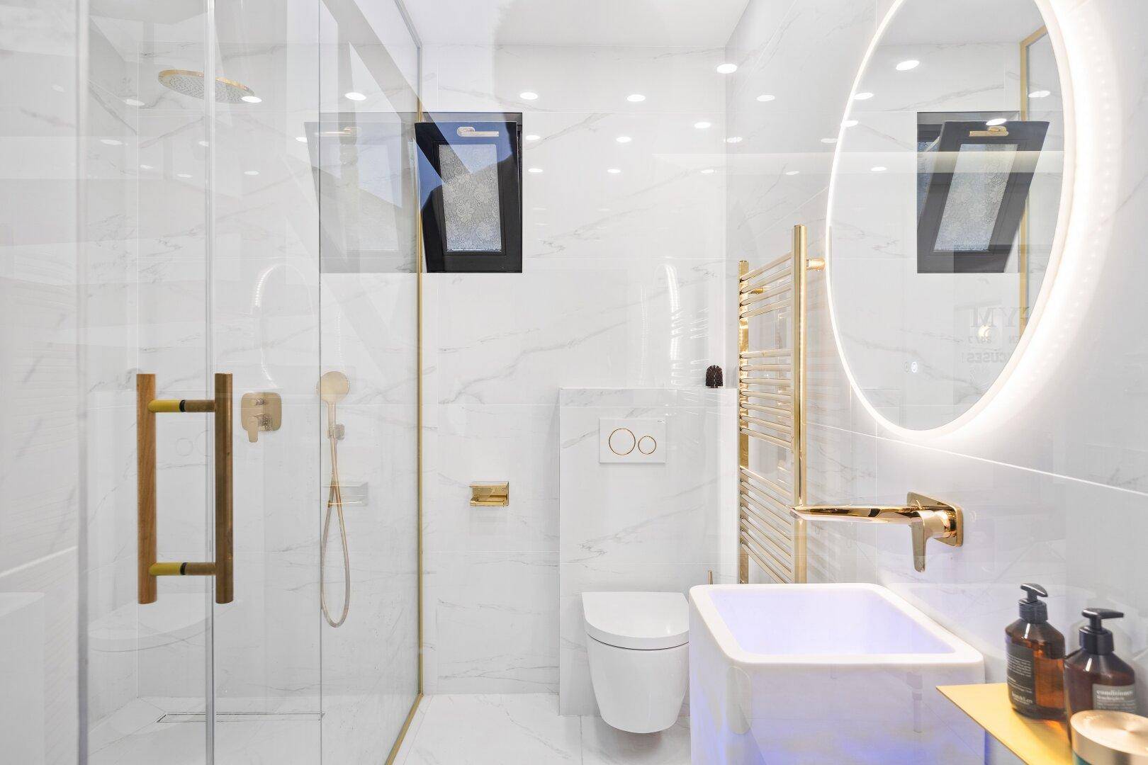 Helles Badezimmer mit Marmorfliesen, Glasdusche und goldfarbenen Armaturen.
