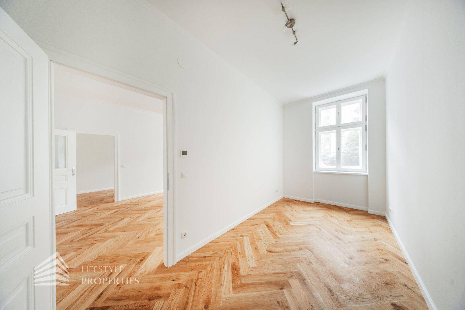 Lichtdurchfluteter Raum mit Fischgrätparkett und Zugang zu weiteren Zimmern.