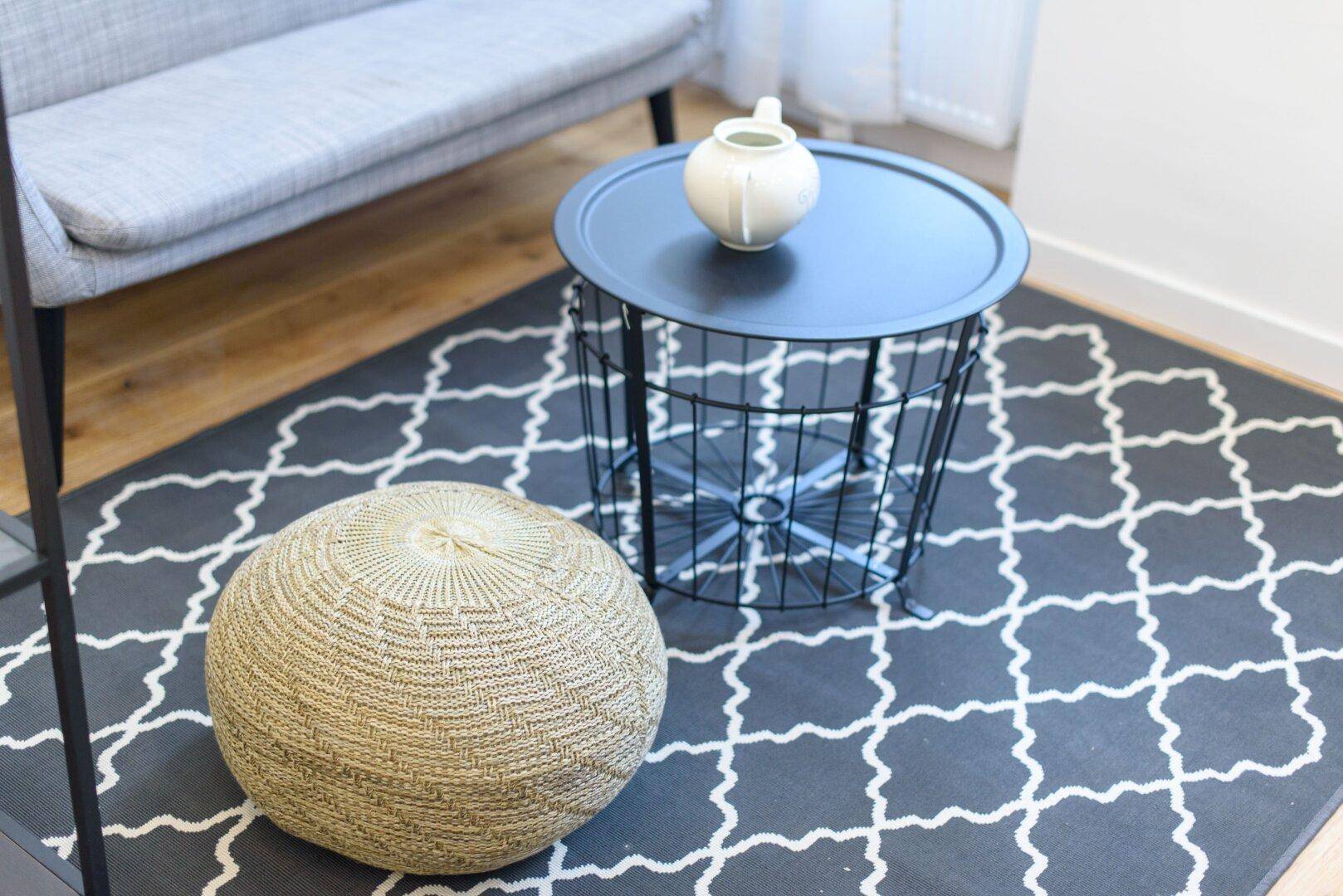 Detailaufnahme des Wohnzimmerbodens mit gemustertem Teppich, Couchtisch und einem gemütlichen Pouf.