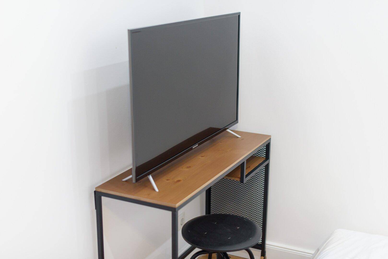 Flacher Fernseher auf einem schmalen Schreibtisch mit Metallgestell und einem passenden Hocker.
