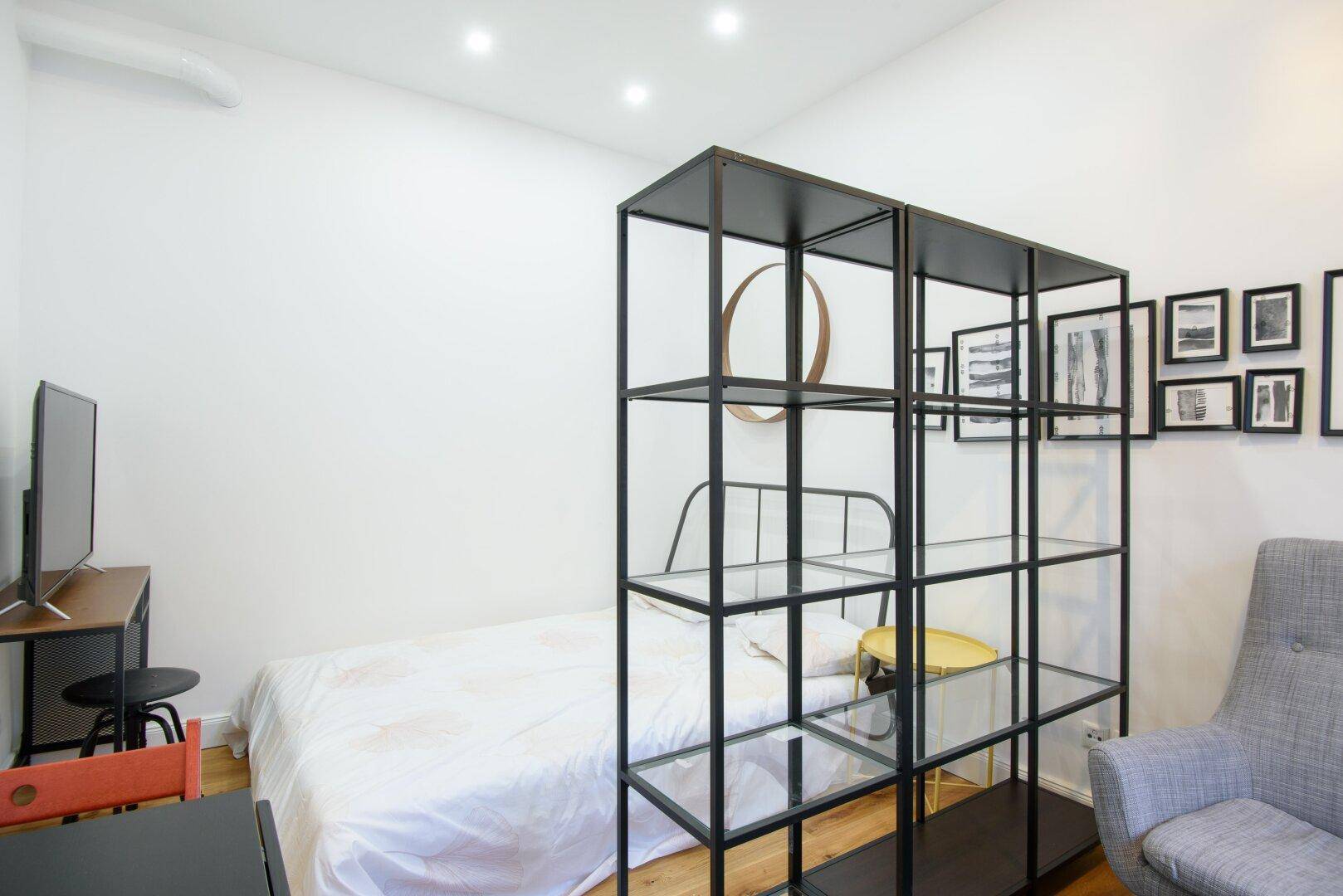 Schlafzimmer mit Bett, TV-Bereich und einem Regal als Raumteiler in modernem Design.