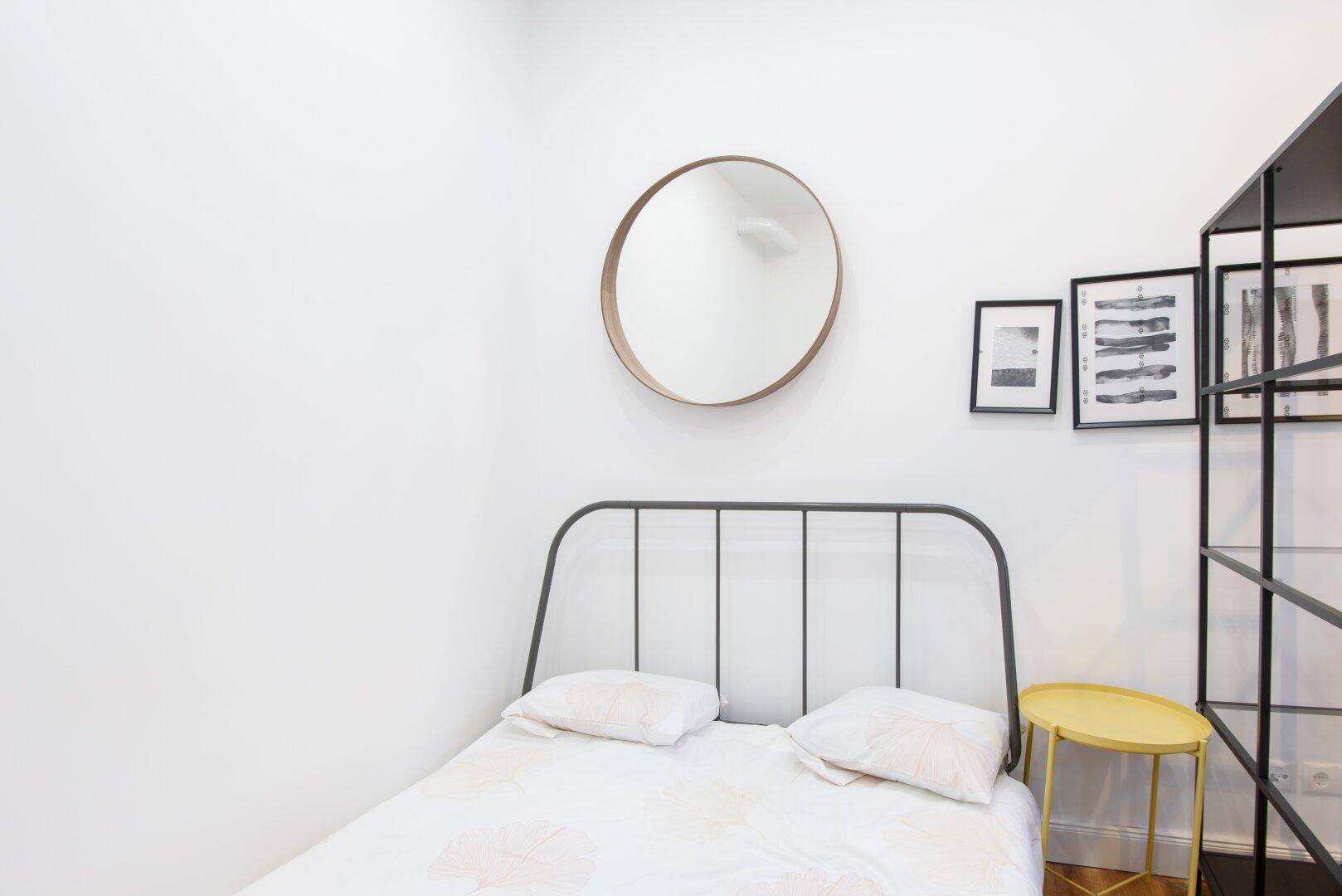 Modernes Schlafzimmer mit minimalistischem Bettgestell, rundem Spiegel und hellem Beistelltisch.