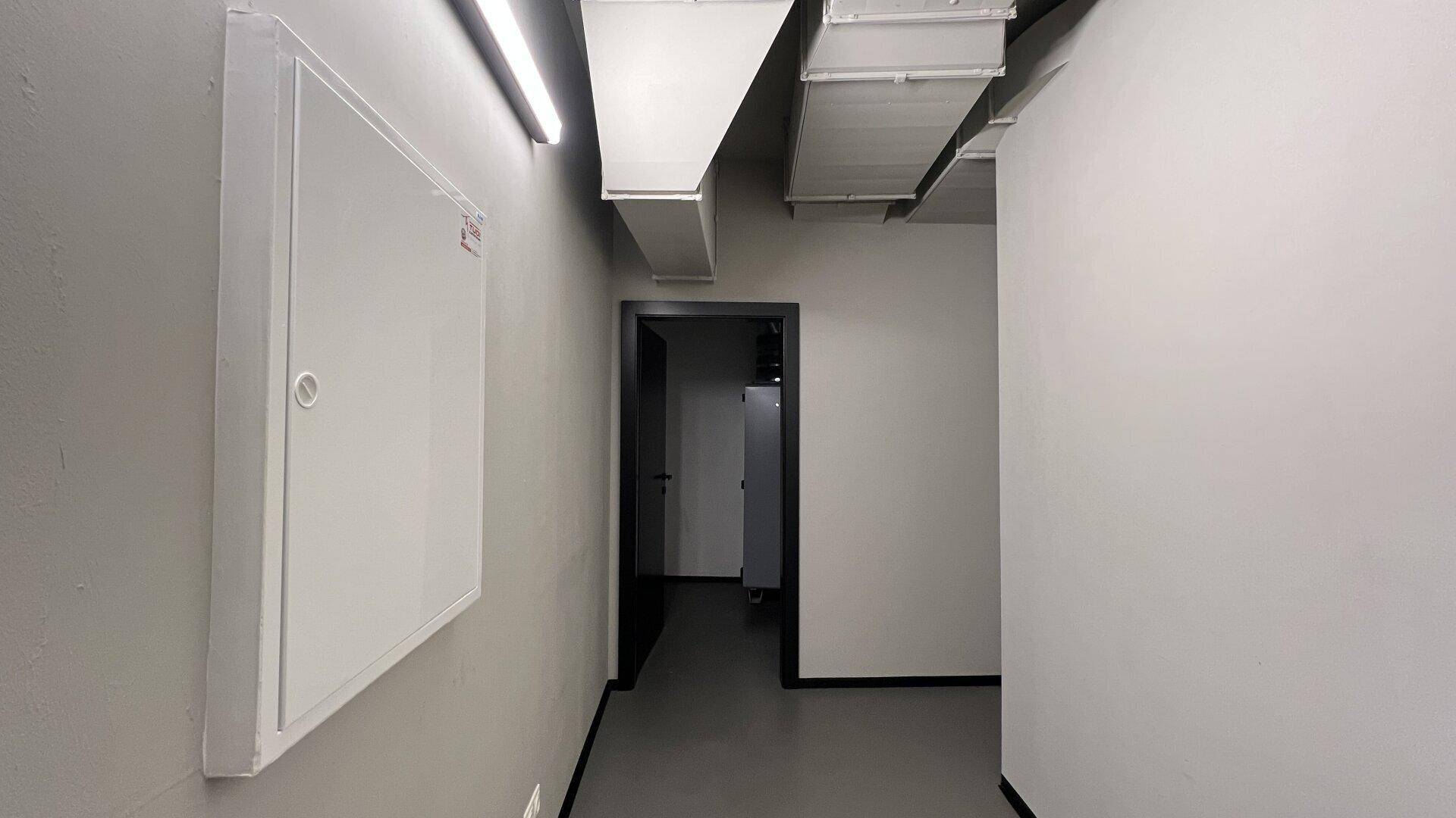 Schmaler Flur mit weißen Wänden, industrieller Deckenbeleuchtung und sichtbaren Lüftungskanälen.