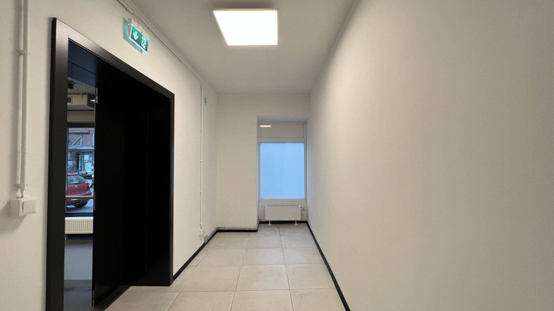 Heller Flur mit hellgrauen Fliesen, weißen Wänden und einem Fenster am Ende, inklusive Aufzugzugang.