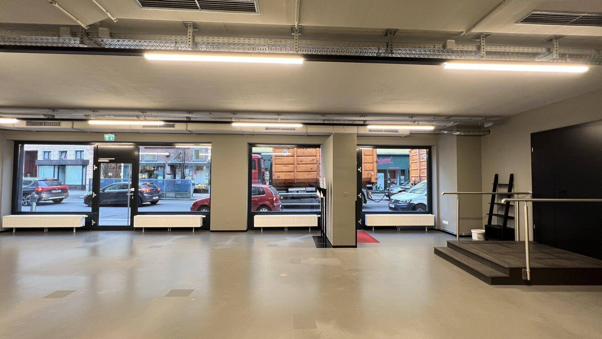 Weitläufiger Geschäftsraum mit großen Schaufenstern, die viel Tageslicht hereinlassen und eine kleine erhöhte Plattform.