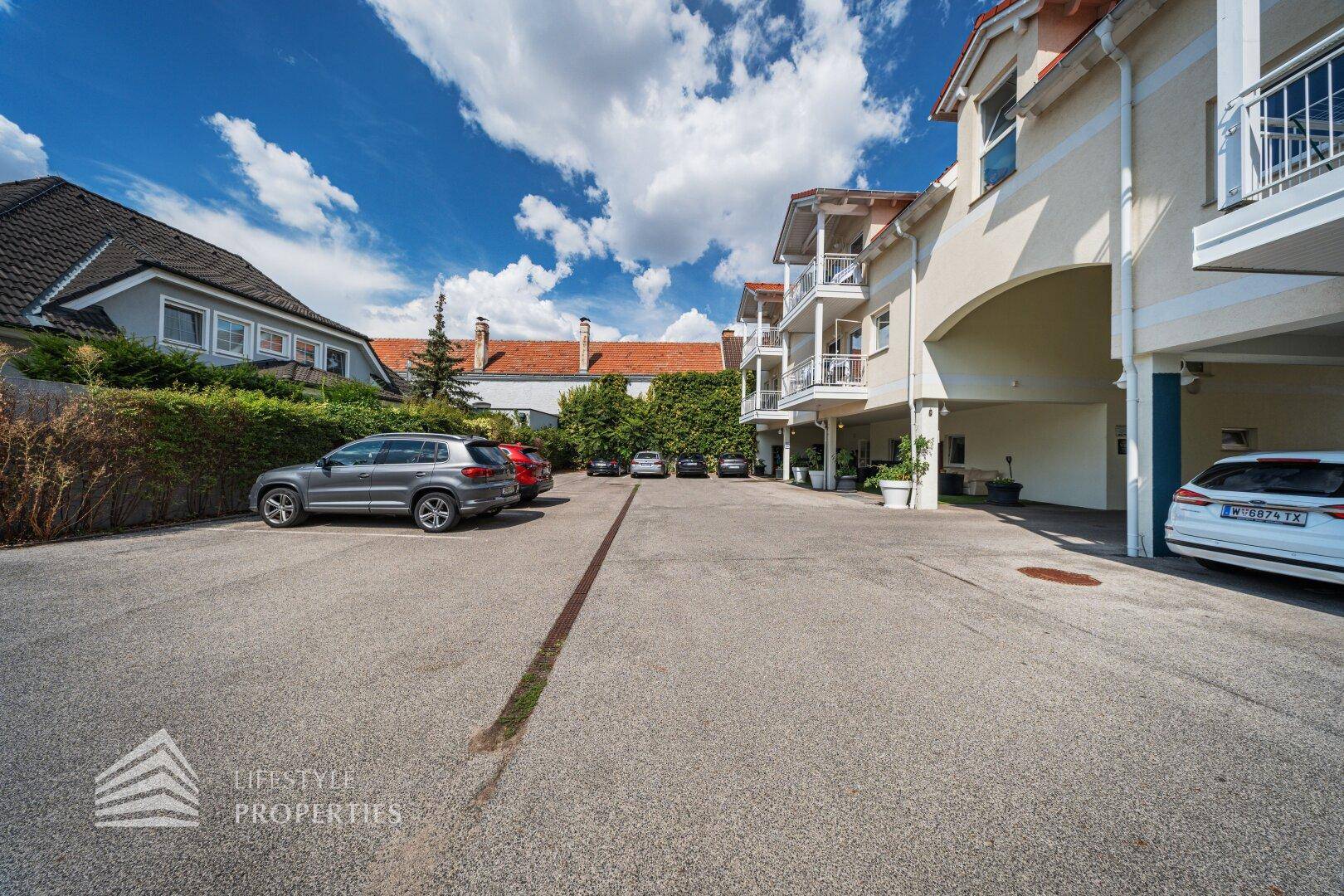 Großer Außenparkplatz mit mehreren Fahrzeugen und umgebender Hecke unter blauem Himmel.