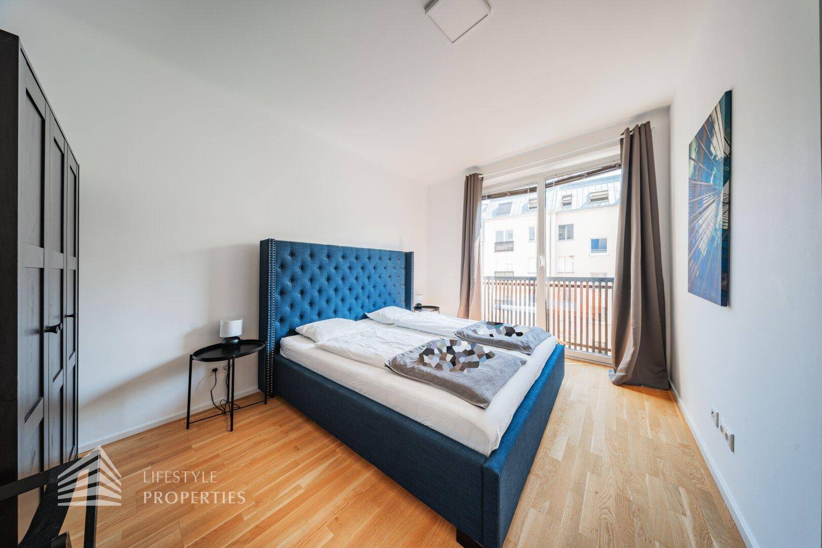 Schlafzimmer mit großem blauen Polsterbett, Holzboden und Zugang zu einem französischen Balkon.