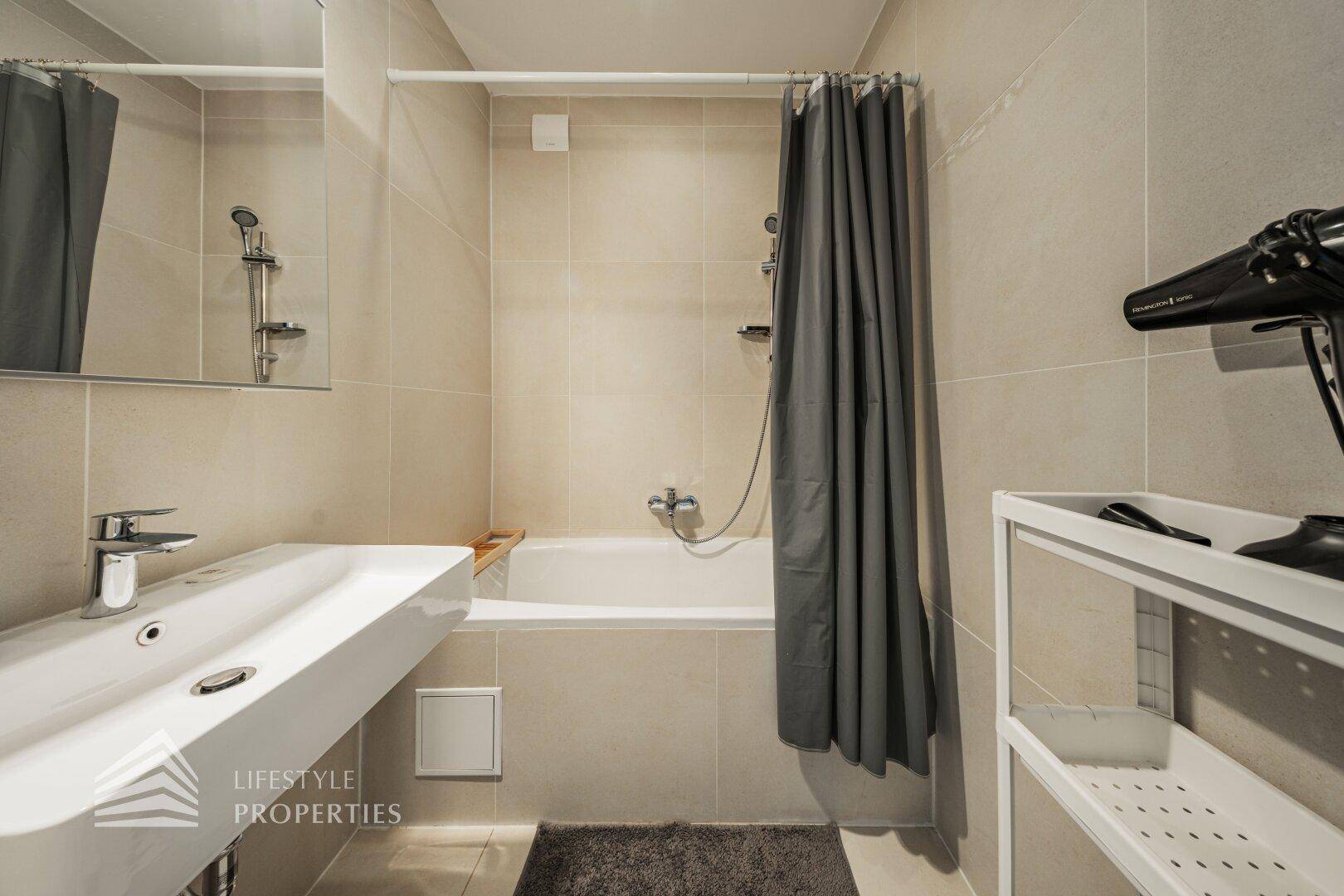 Modernes Badezimmer mit beigefarbenen Fliesen, Badewanne mit Duschvorhang und elegantem Waschbecken.
