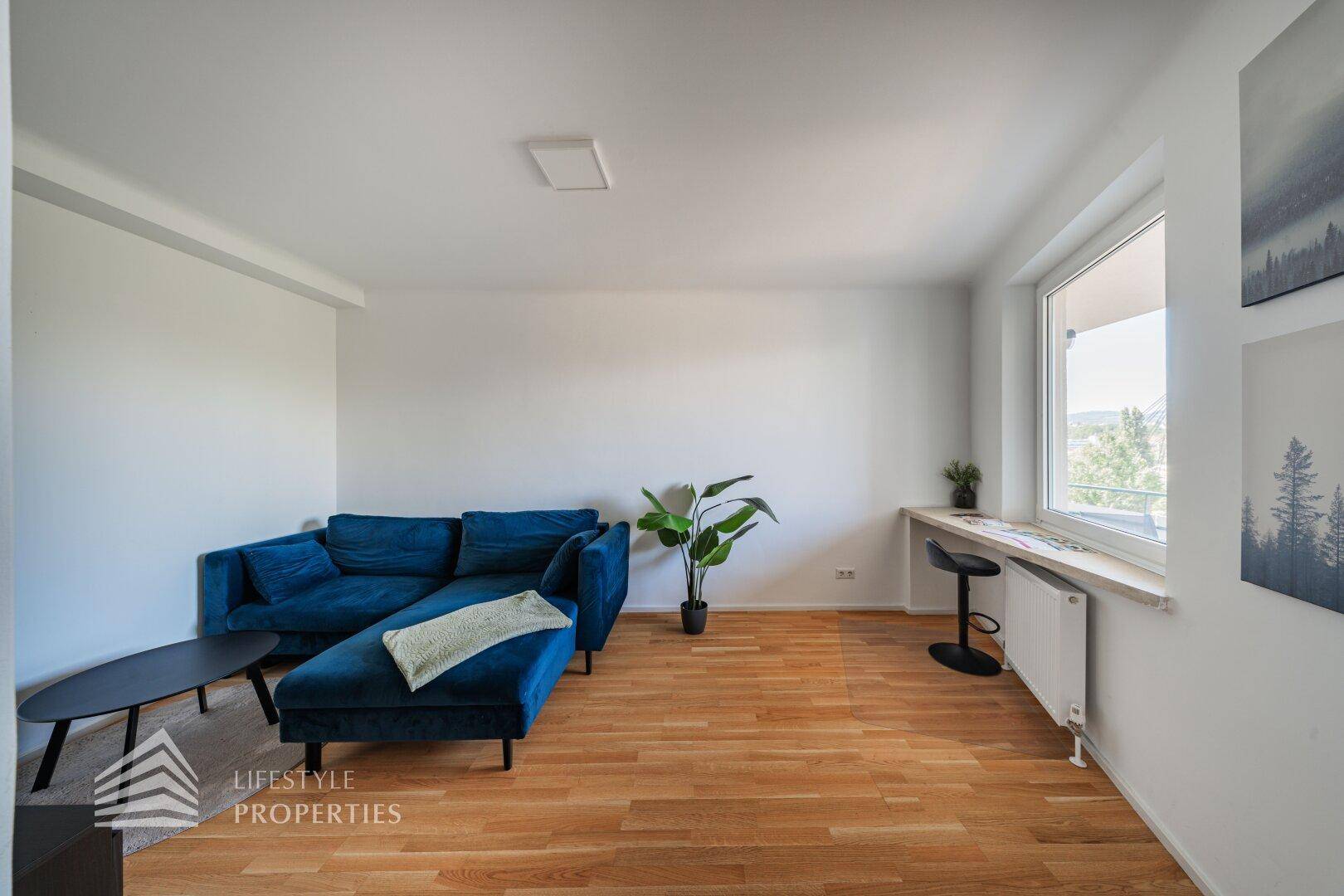Gemütliches Wohnzimmer mit blauer Chaiselongue, Holzboden und einem Arbeitsplatz am Fenster.