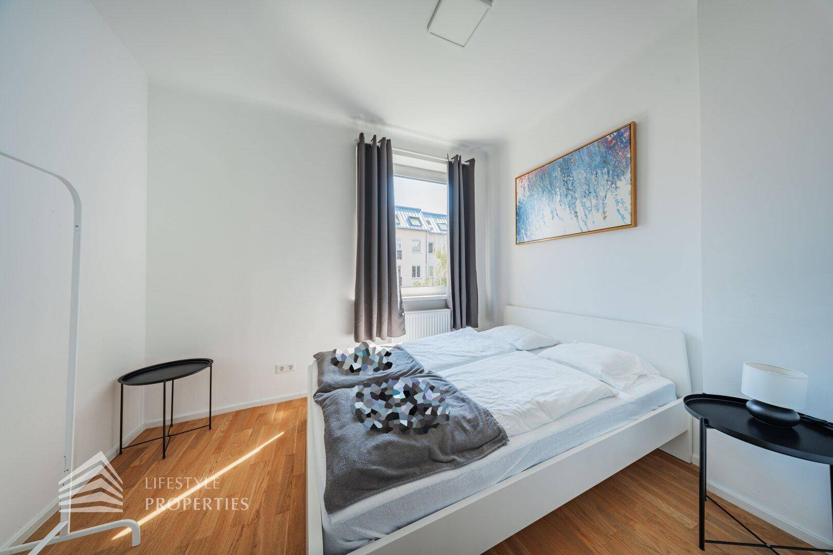 Helles Schlafzimmer mit weißem Bett, Holzboden und einem Fenster, das viel Tageslicht hereinlässt.