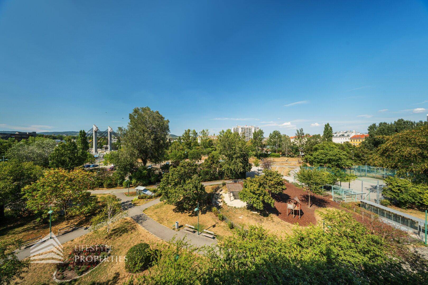 Weitläufiger Blick auf eine grüne Parkanlage mit Bäumen, Wegen und einem Spielplatz.