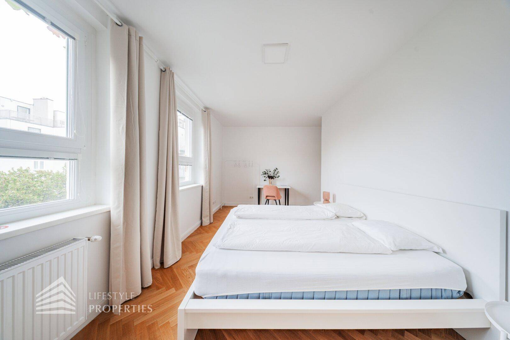 Helles Schlafzimmer mit Doppelbett, Fischgrätparkett und großen Fenstern.