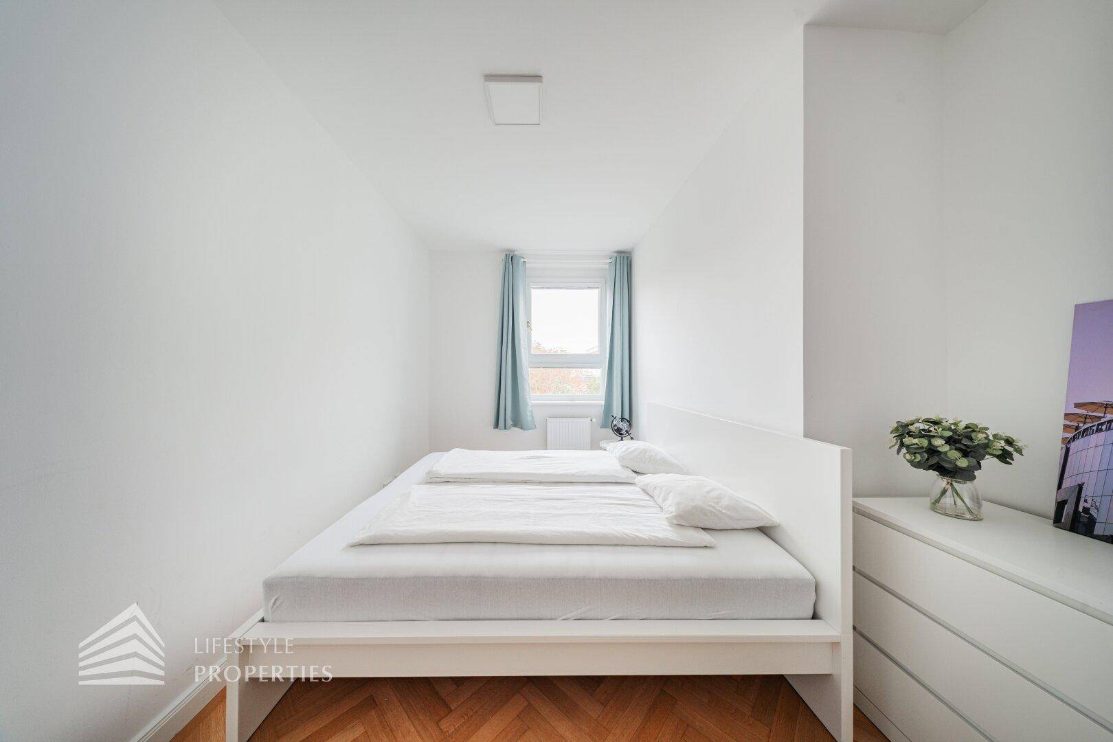 Gemütliches Schlafzimmer mit Doppelbett, weißer Kommode und Fischgrätparkett.