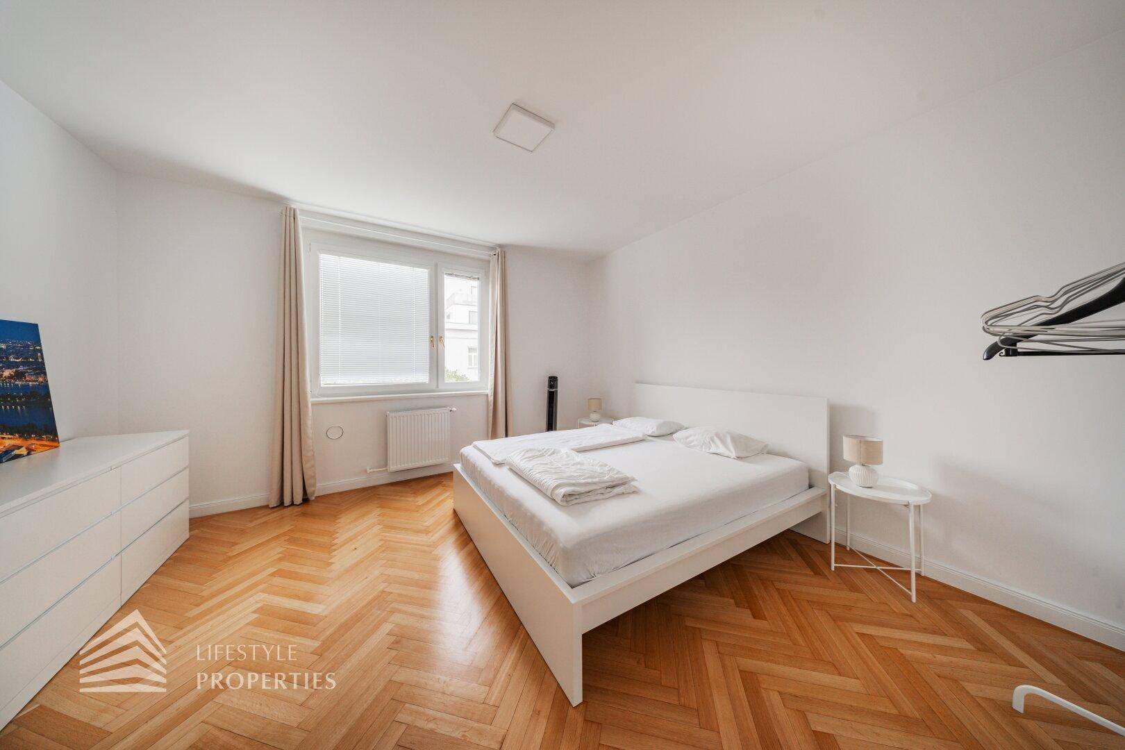 Helles Schlafzimmer mit Fischgrätparkett, Doppelbett und Fenster mit Blick nach draußen.