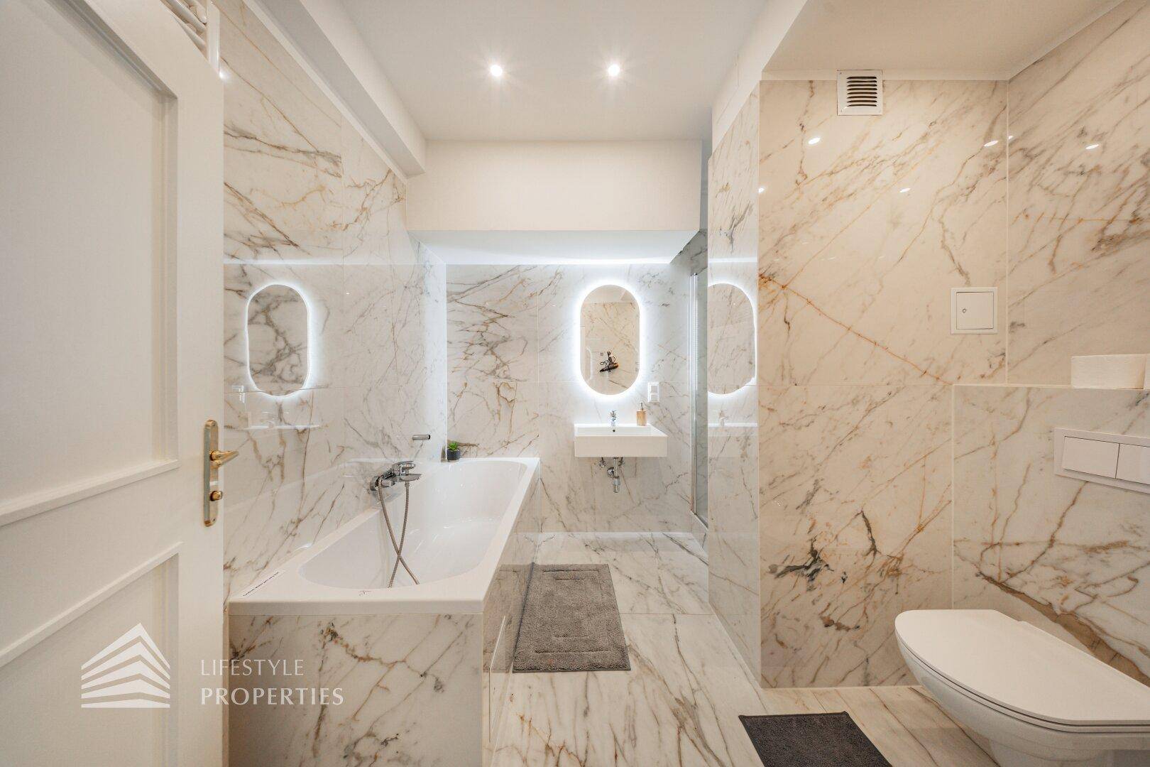 Modernes Badezimmer mit Marmorfliesen, Badewanne und beleuchtetem Spiegel.