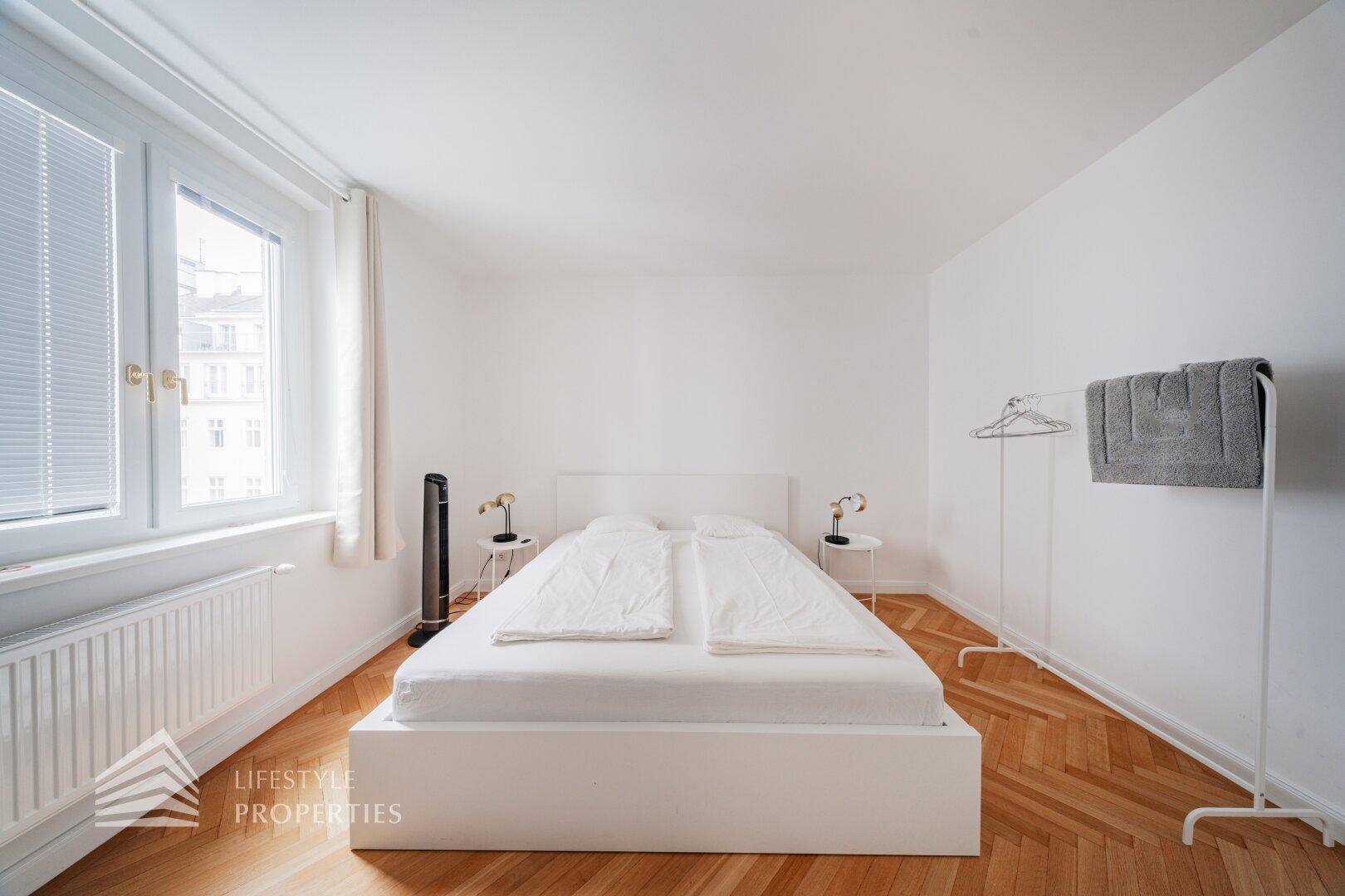 Helles Schlafzimmer mit Doppelbett, Fischgrätparkett und großem Fenster.