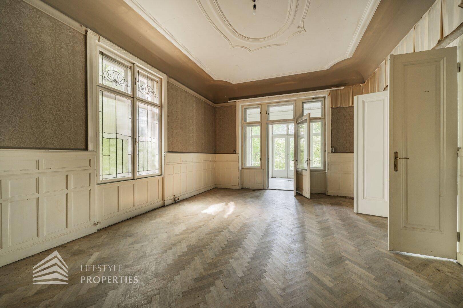 Geräumiges Wohnzimmer mit Fischgrätparkett, hohen Decken und dekorativen Wandpaneelen, bereit zur Renovierung.