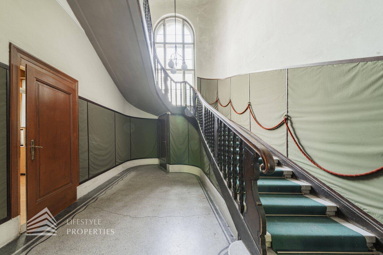 Repräsentatives Treppenhaus mit breiter Holztreppe, grünem Teppich und großem Bogenfenster.