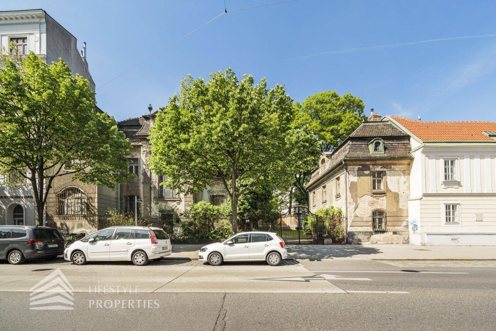 Historische Gebäude entlang einer städtischen Straße mit parkenden Autos und viel Grün.