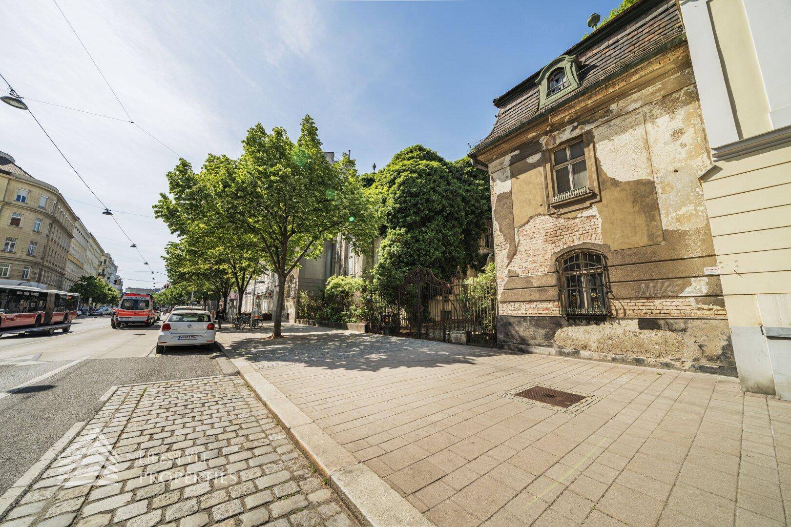 Historische Fassade eines Gebäudes an einer belebten Straße mit Kopfsteinpflaster und Bäumen.