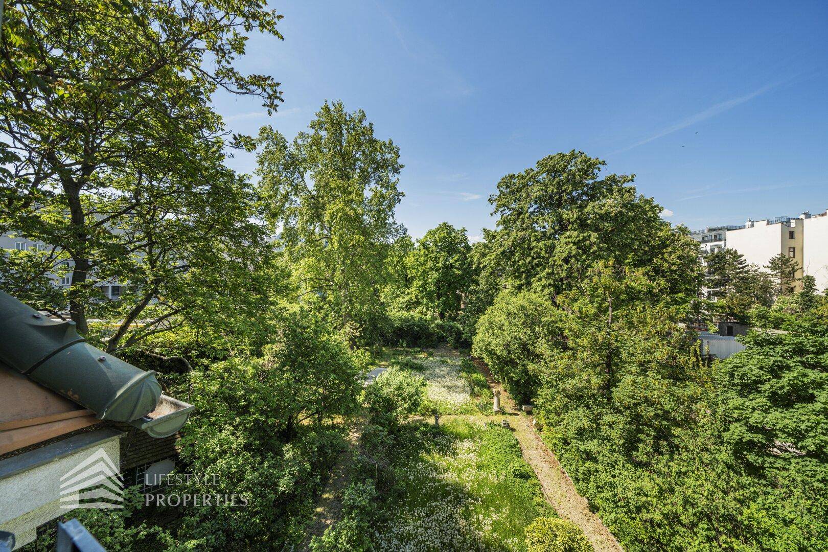Weitläufiger Blick über einen grünen Garten mit hohen Bäumen und umliegenden Gebäuden.
