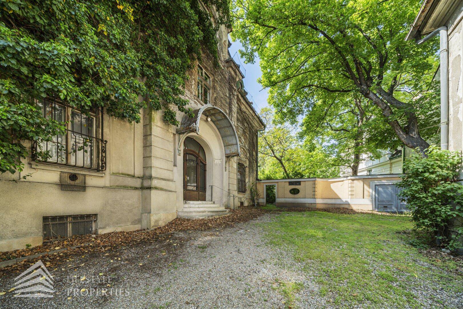 Seitlicher Eingang der Villa mit Kiesweg und bewachsenen Mauern, umgeben von altem Baumbestand.