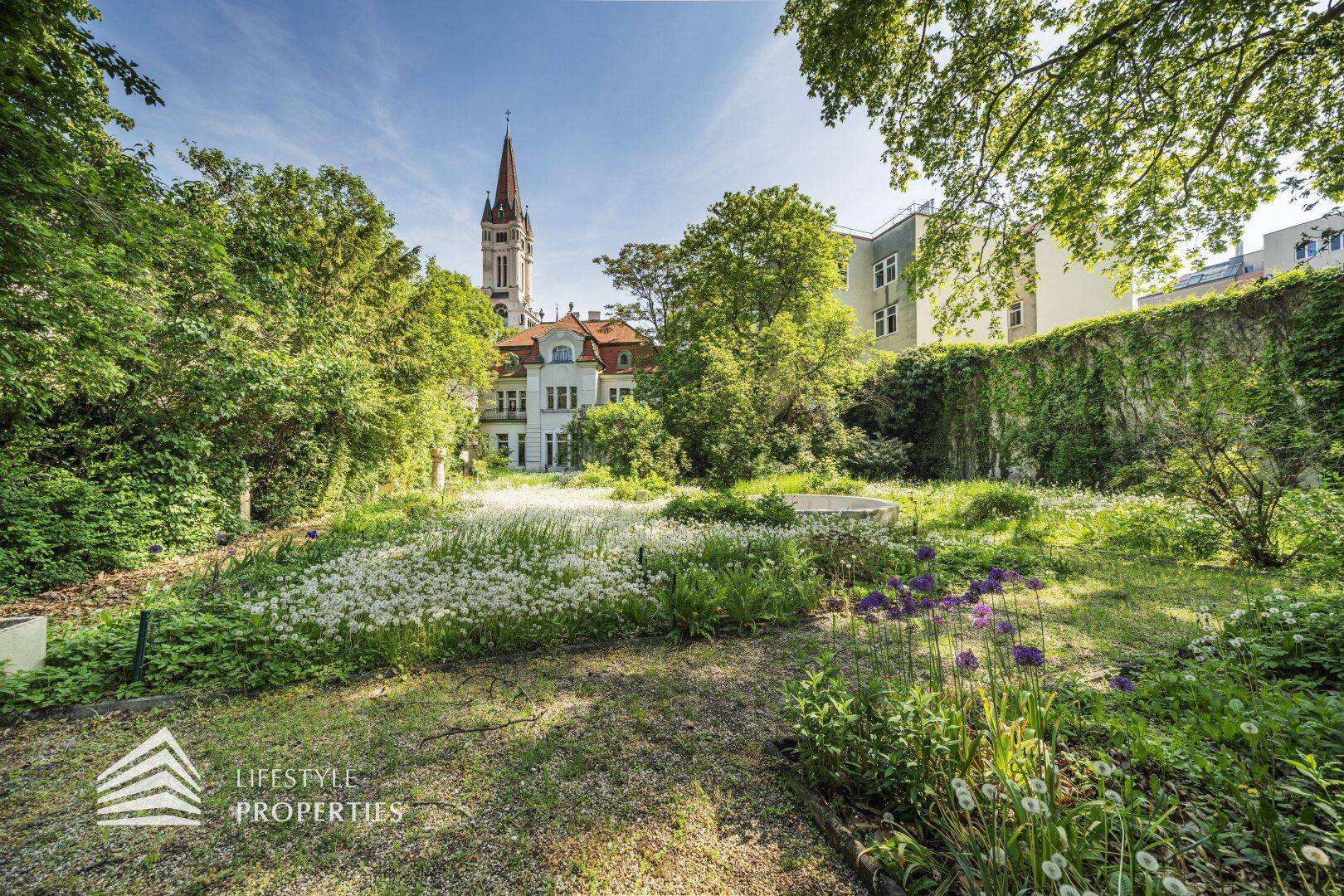 Großer, gepflegter Garten mit blühenden Pflanzen und Blick auf die historische Villa und Kirchturmspitze.