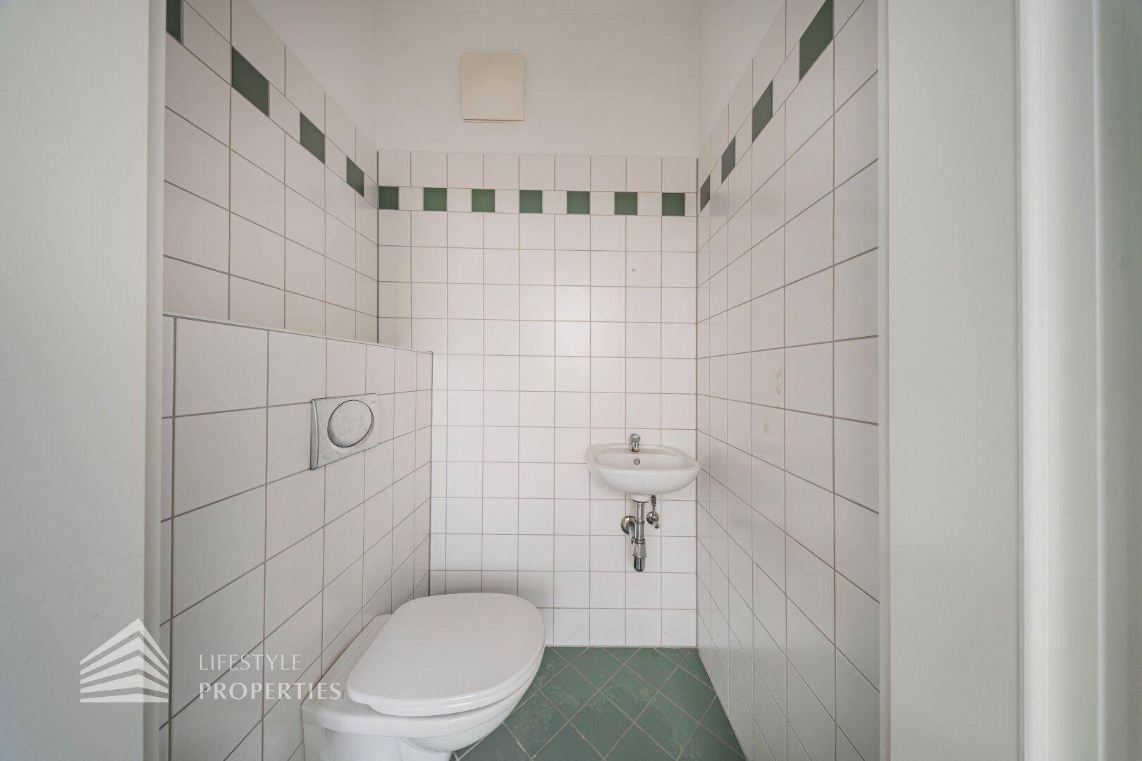 Separate Gästetoilette mit WC, kleinem Waschbecken und weißen Fliesen mit grünen Akzenten.