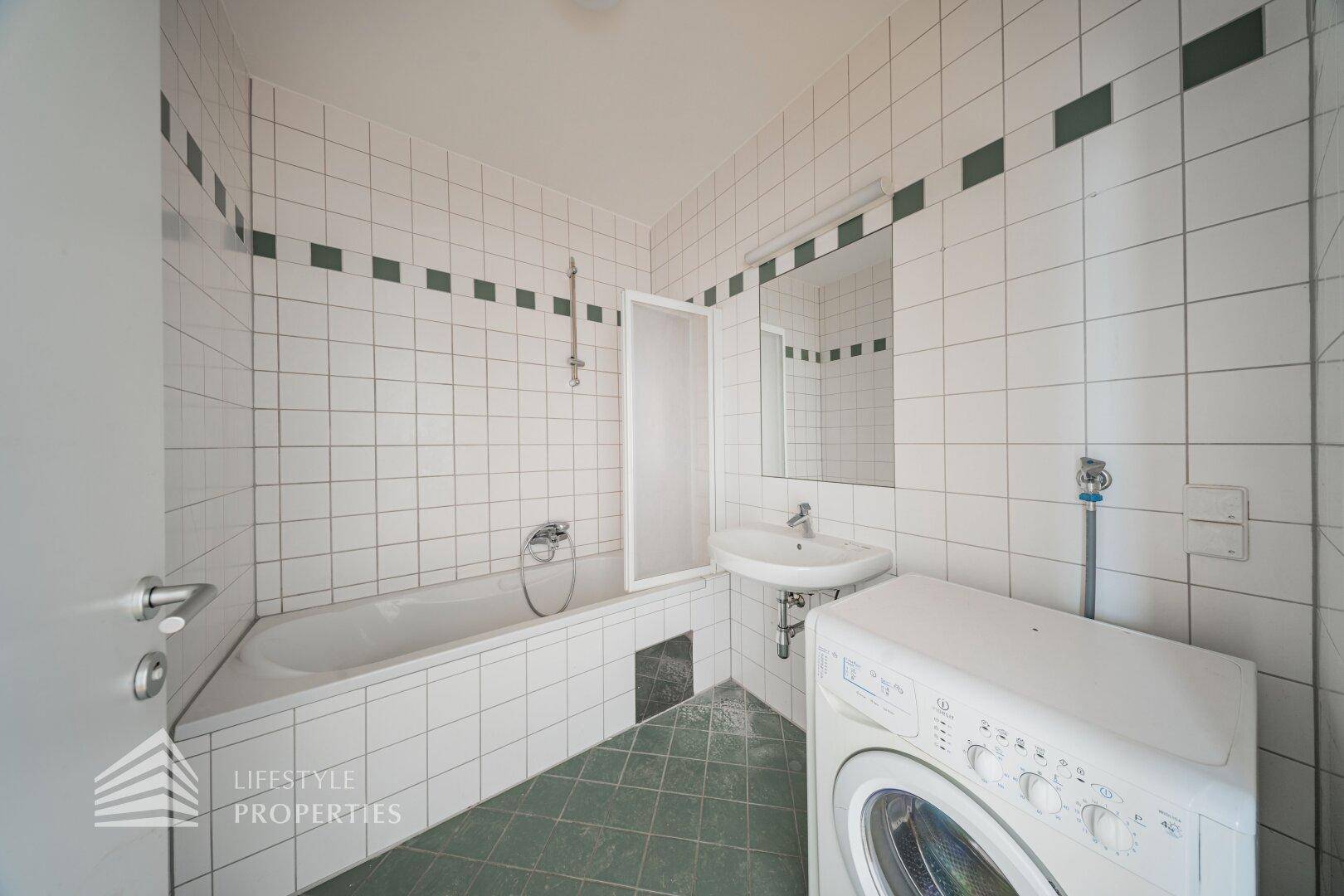 Badezimmer mit Badewanne, Waschbecken und Waschmaschinenanschluss, gefliest in Weiß und Grün.