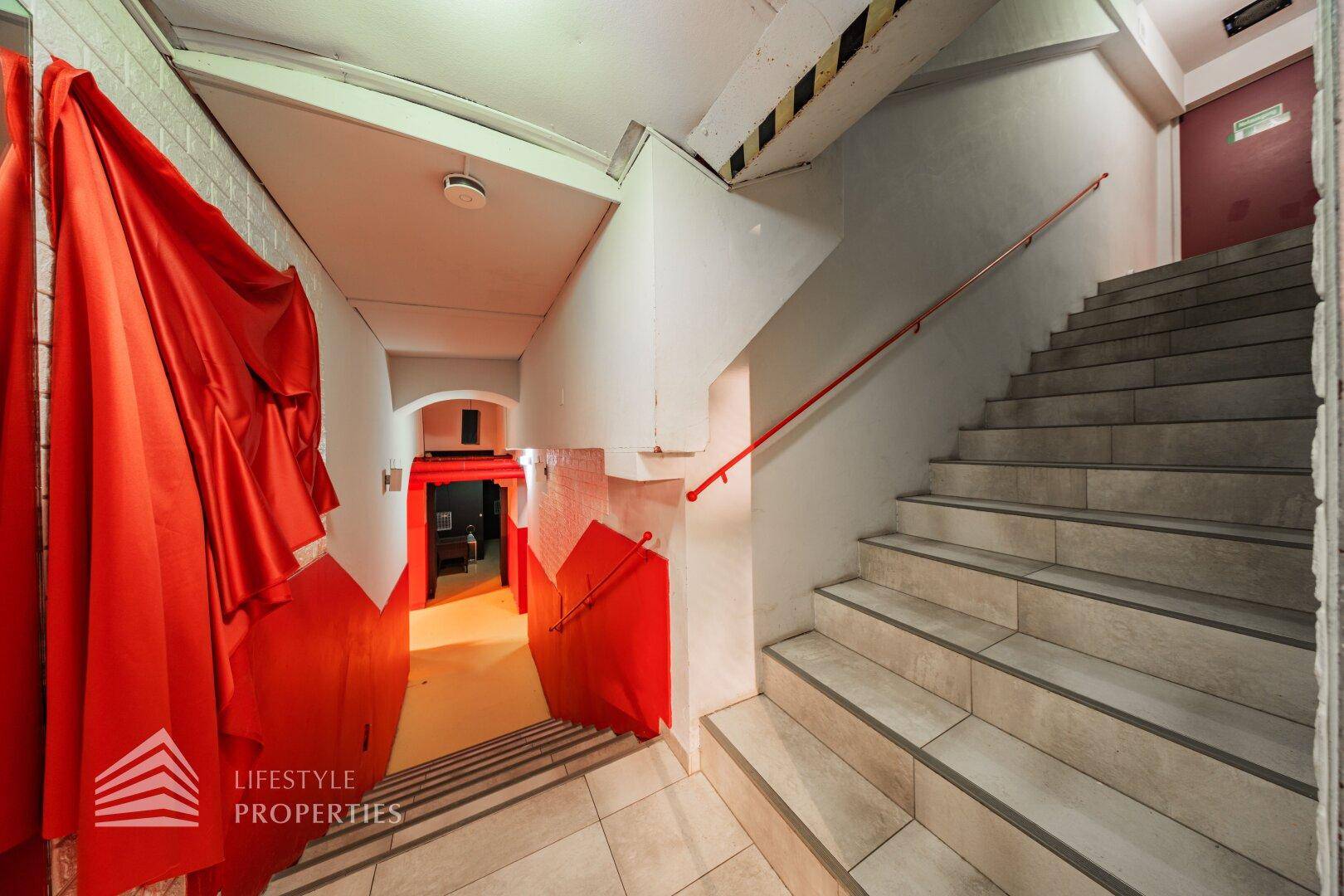 Treppenhaus mit gefliesten Stufen, rotem Handlauf und weiß gestrichenen Wänden, die nach unten führen.