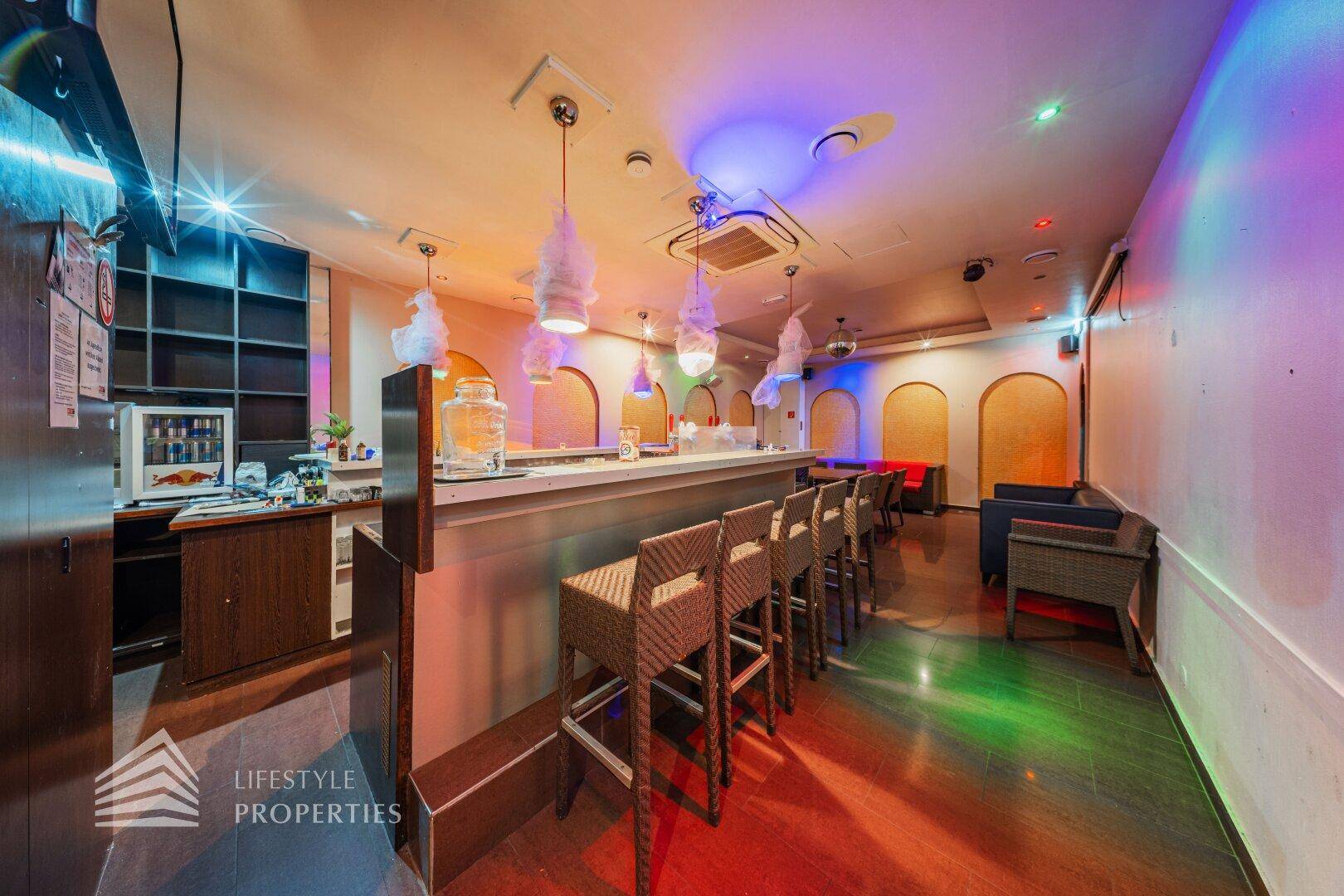 Bar-Bereich mit Theke, Barhockern und Lounge-Sitzgelegenheiten, beleuchtet von farbigen Lichtern.