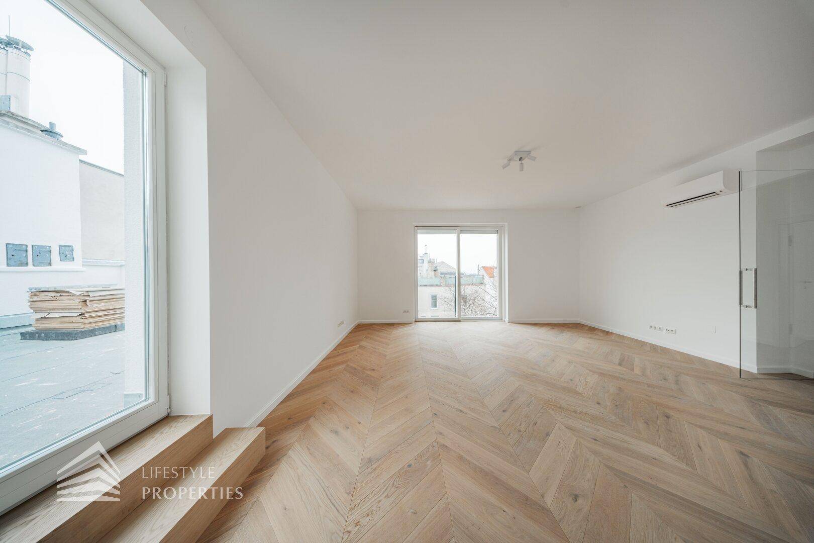 Geräumiges, helles Zimmer mit Fischgrätparkett und großen Fenstern, die viel Tageslicht hereinlassen.