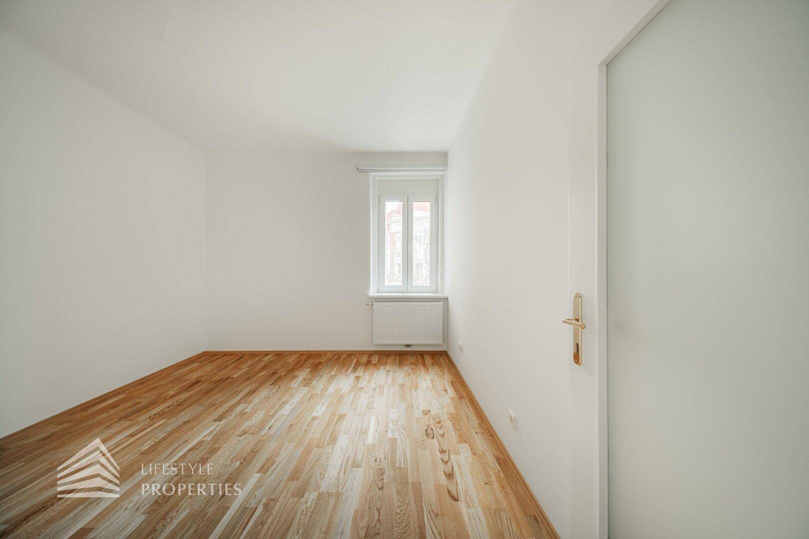 Helles Schlafzimmer mit Parkettboden und einem Fenster mit Rollladen.