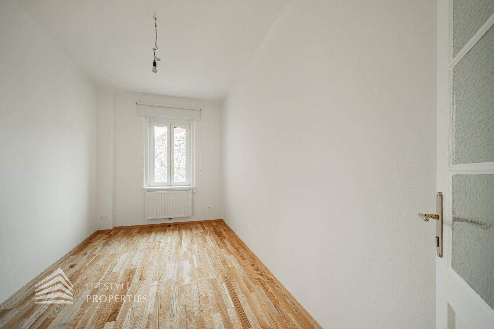 Kleines, helles Zimmer mit Parkettboden und einem Fenster mit Rollladen.