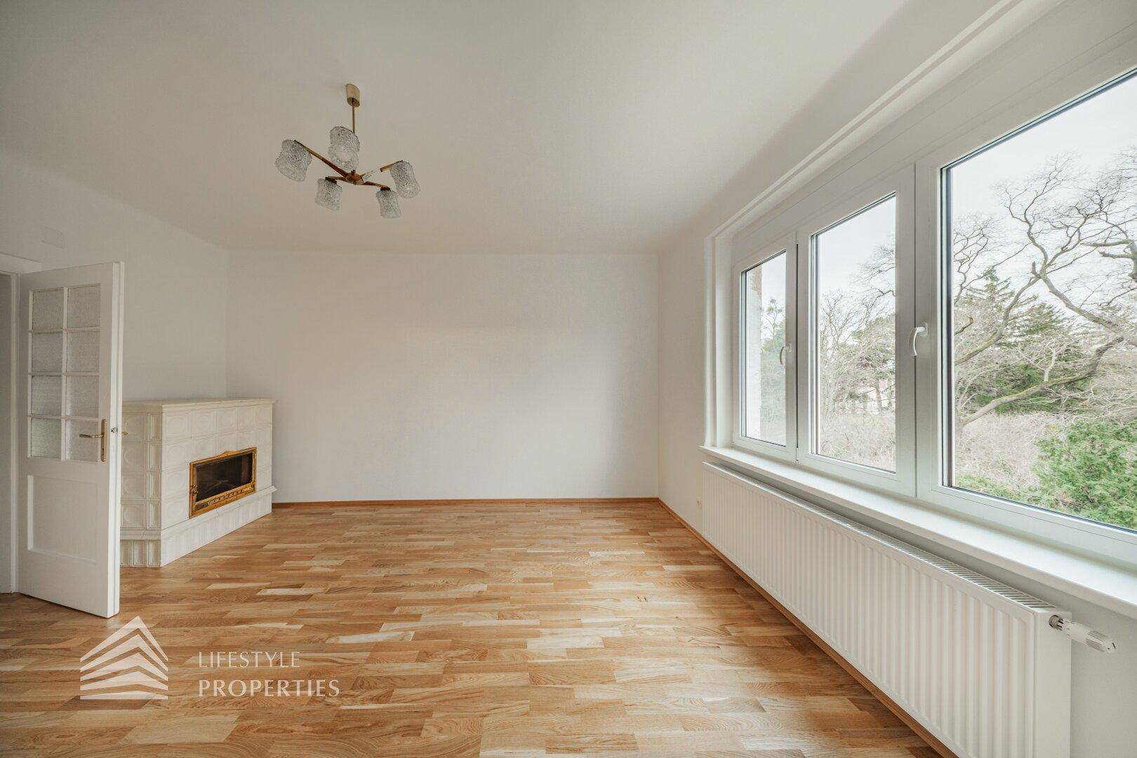 Geräumiges Wohnzimmer mit Parkettboden, Kamin und großen Fenstern mit Blick ins Grüne.