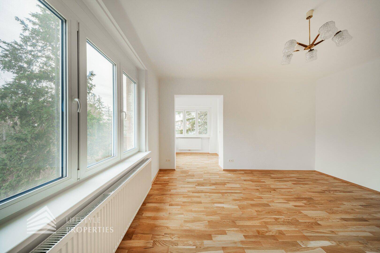 Geräumiges Wohnzimmer mit Parkettboden und großen Fenstern, die eine grüne Aussicht bieten.