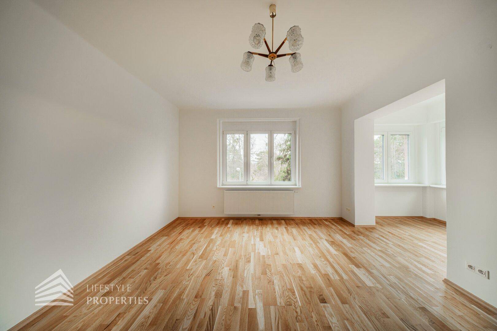 Großes Wohnzimmer mit Parkettboden und zwei Fensterbereichen, die viel Licht spenden.