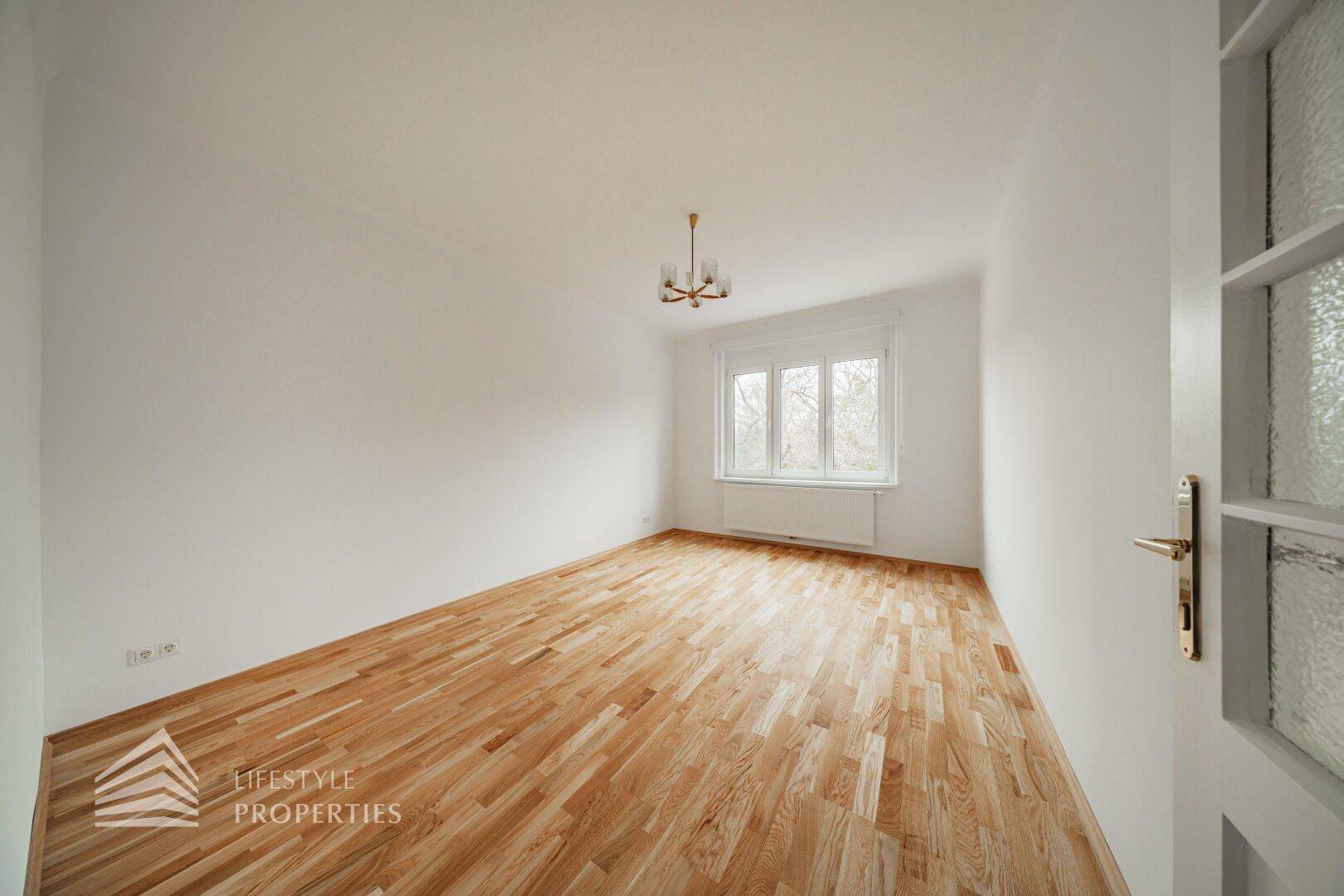 Geräumiges Wohnzimmer mit Parkettboden und einem großen Fenster, das viel Licht hereinlässt.